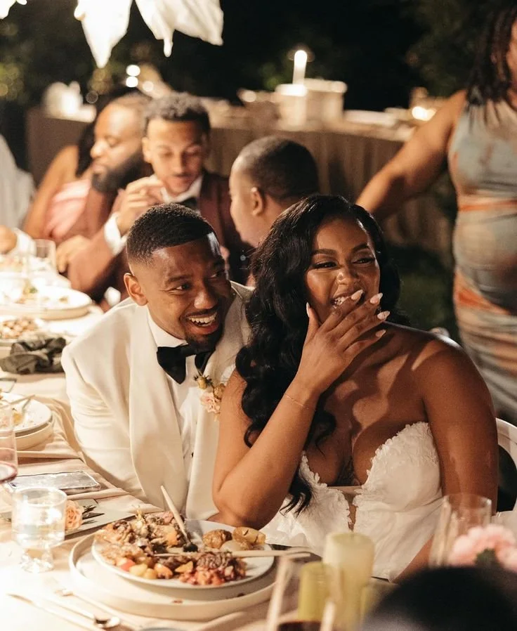 A bride and groom at their wedding reception, laughing and enjoying the moment, seated at a decorated table with food and drinks.