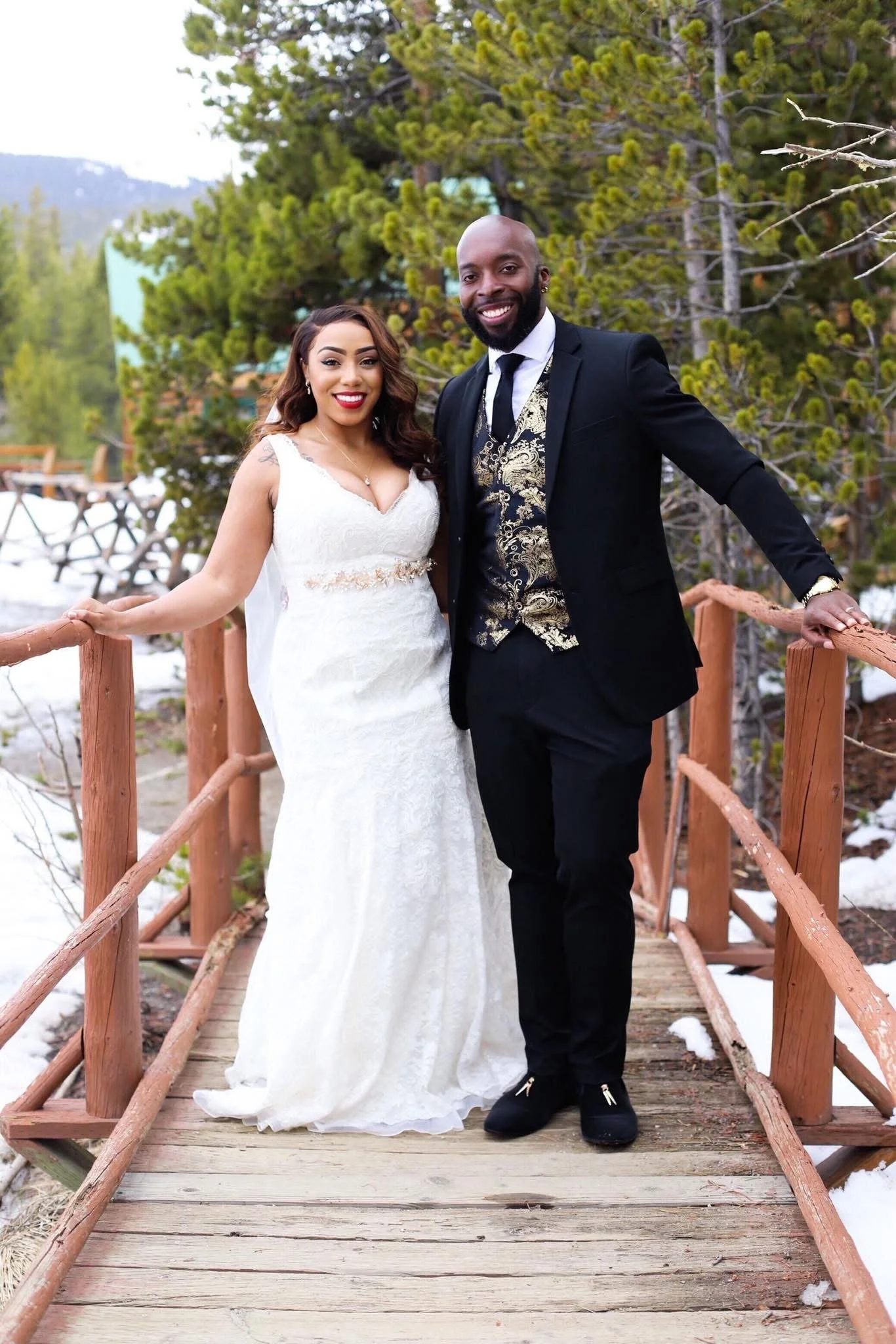 A newlywed couple standing on a small wooden bridge outdoors, with trees and snow in the background. The bride is wearing a white wedding gown and the groom is dressed in a black suit with a gold patterned vest.