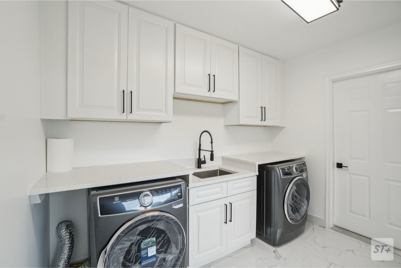 Laundry room with white cabinets, a black countertop, a laundry sink, a black faucet, and two front-loading washing machines. The space has white walls, a white door, and a white marble floor.