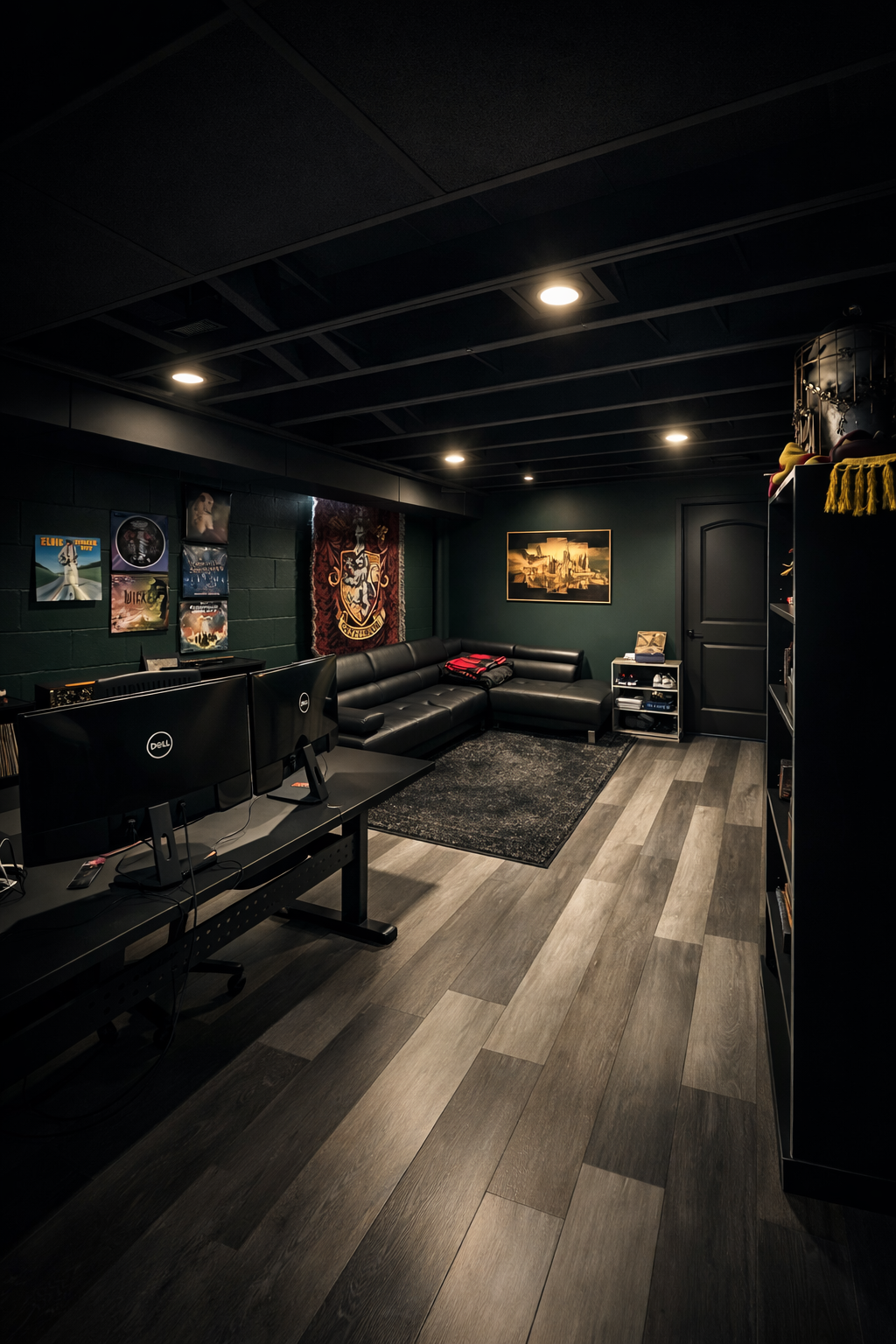 A cozy gaming or entertainment room with black walls, ceiling, and furniture, featuring a black leather sectional sofa, wall art, a bookshelf, a rug, and two computer monitors on a desk.