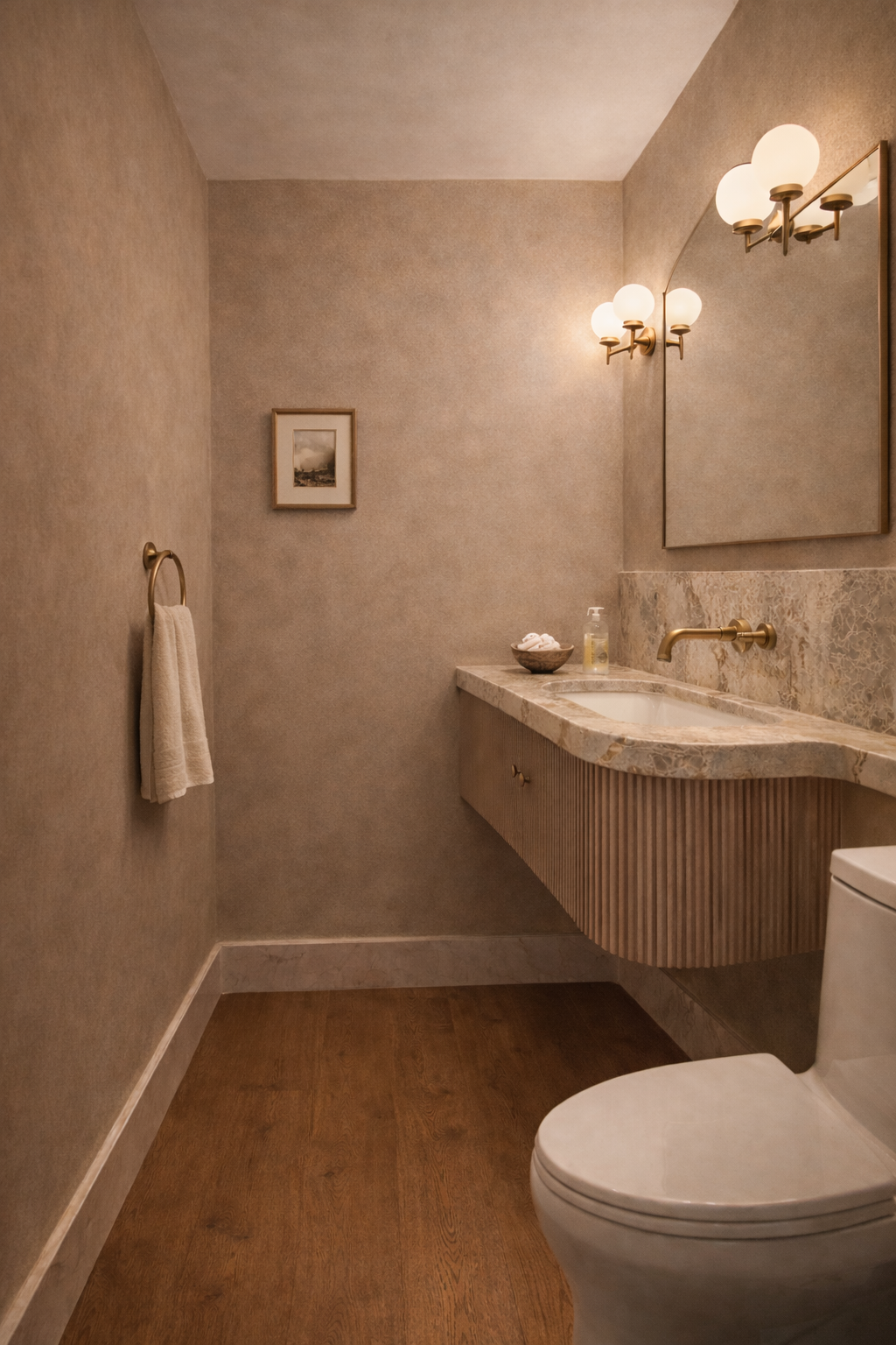 A minimalist bathroom with beige walls, a marble countertop sink, a large mirror, wall-mounted brass light fixtures, a framed picture on the wall, a towel ring with a white towel, and a toilet.