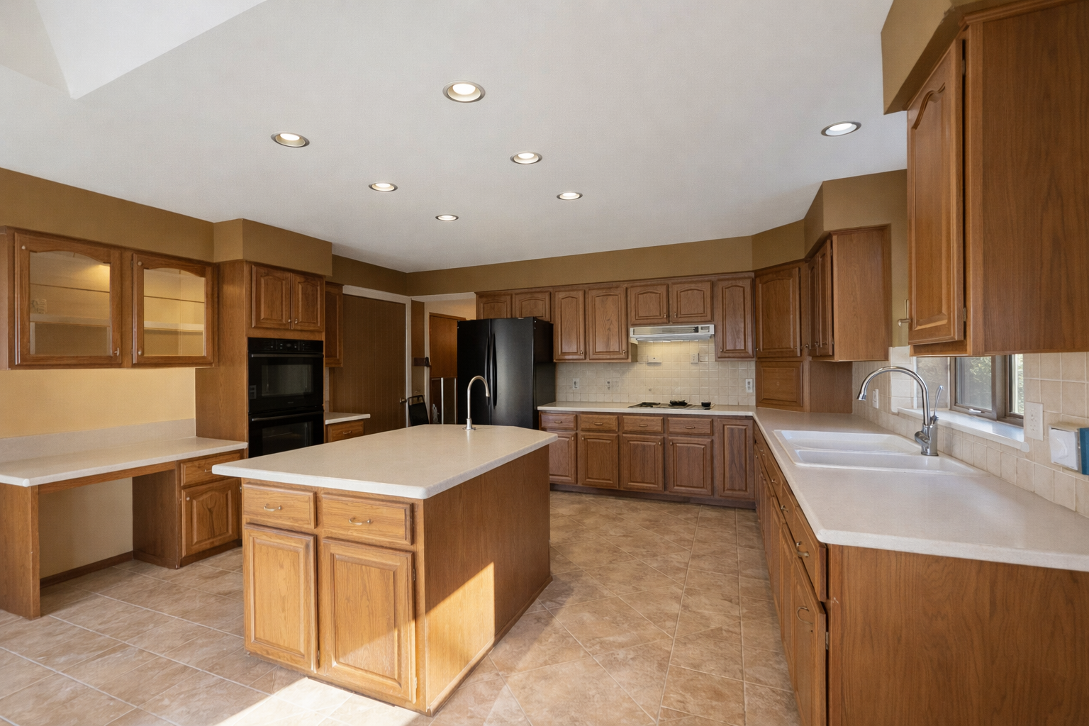 Kitchen with wooden cabinets, beige countertops, a central island with a sink, a black refrigerator, a built-in oven, and a cooktop, with tiled flooring and a window above the sink.