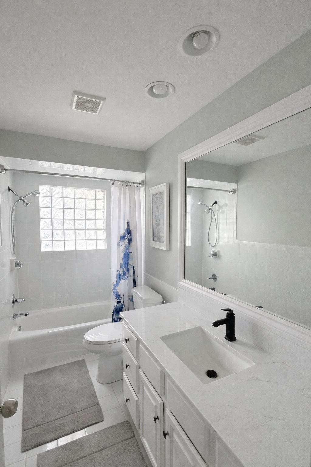 A clean, white bathroom featuring a bathtub with a showerhead, a glass block window, a toilet, a white vanity with a black faucet, a large mirror, and light gray walls.