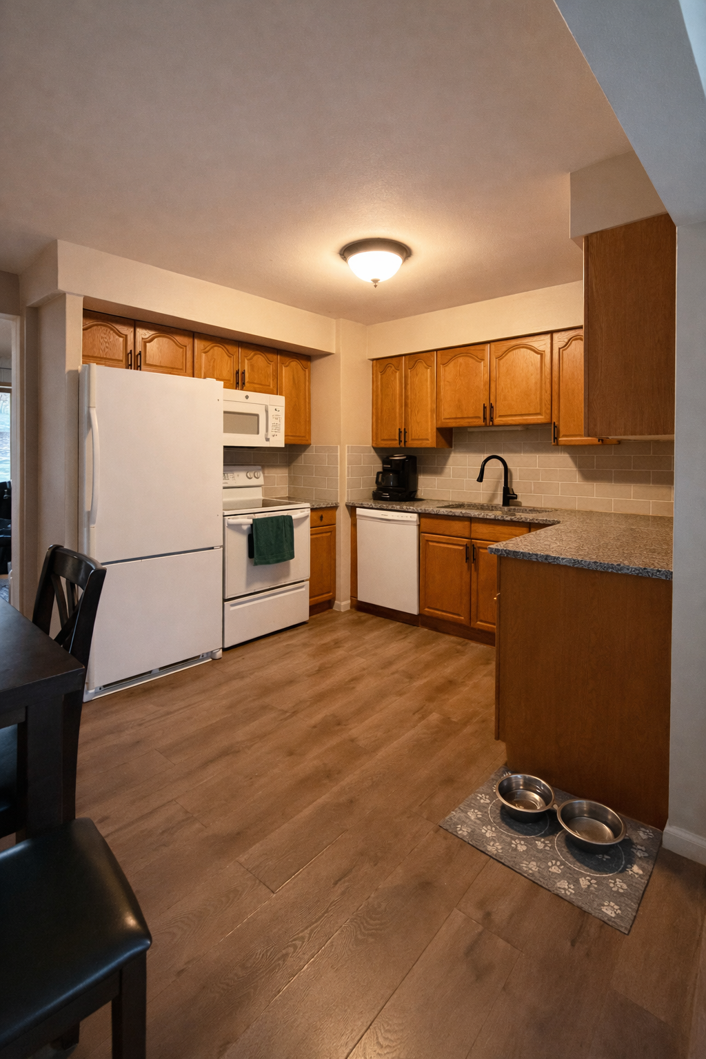 A cozy kitchen with wooden cabinets, white appliances including a refrigerator, microwave, stove, and dishwasher, a black faucet, granite countertops, and two pet bowls on a mat.