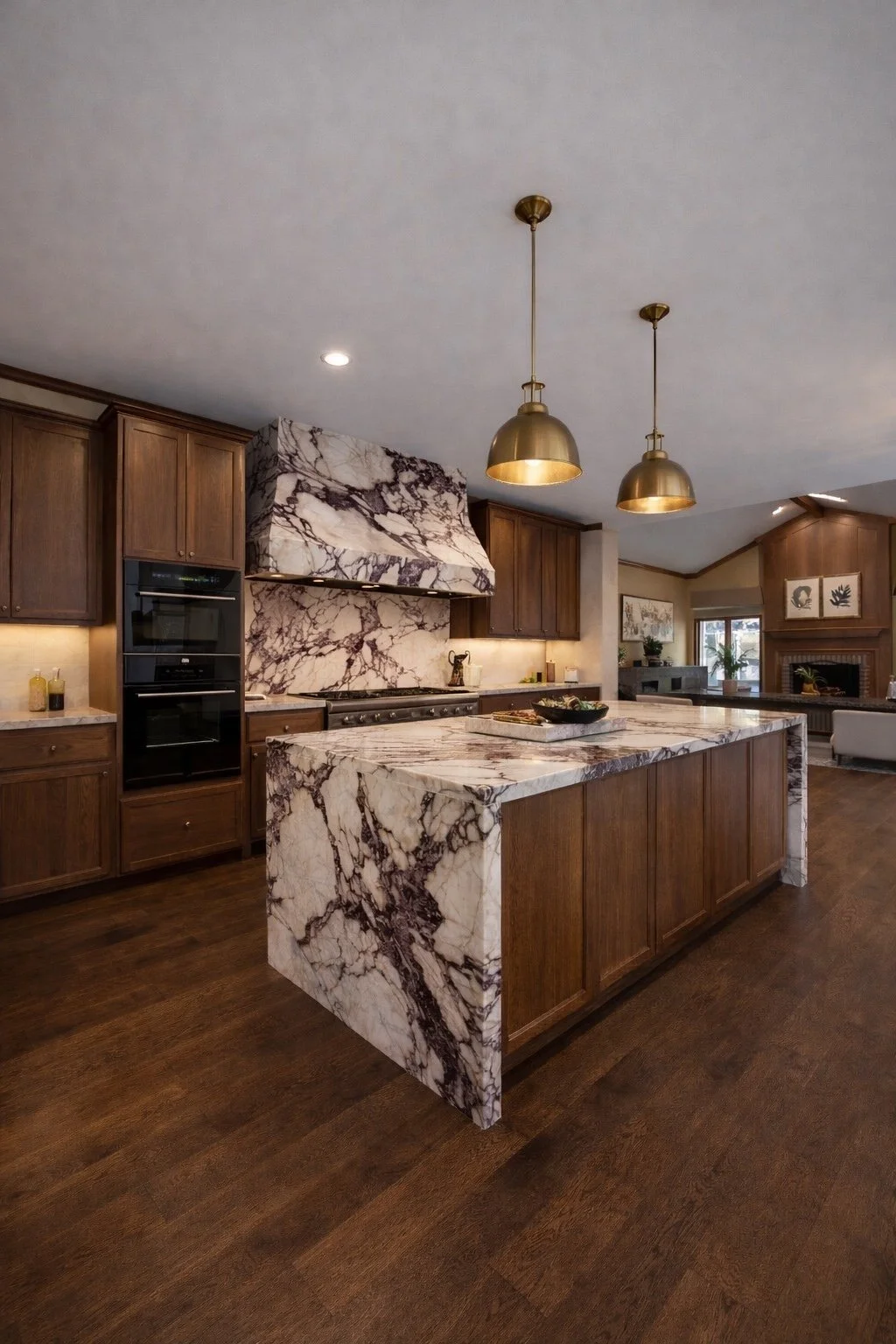 Modern kitchen with wooden cabinets, marble island and backsplash, gold pendant lights, and hardwood flooring.