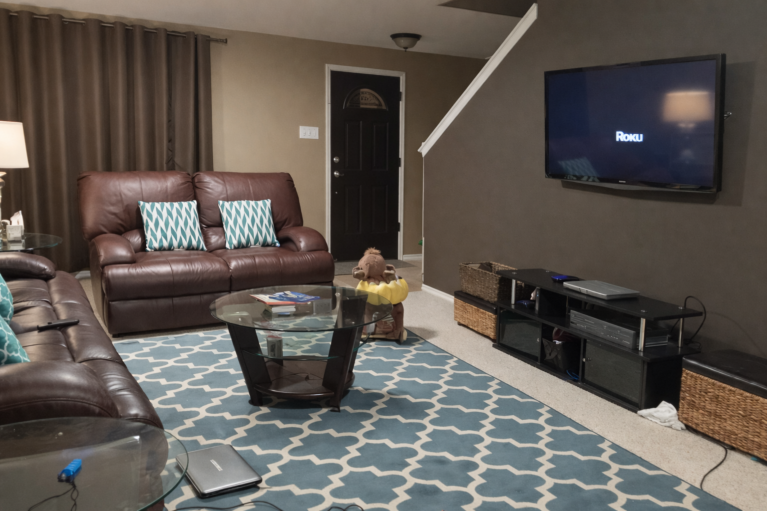 Living room with brown leather sofas with patterned pillows, a coffee table, a blue and white patterned rug, a TV mounted on the wall displaying 'Roku', and various electronics and baskets on a black TV stand.