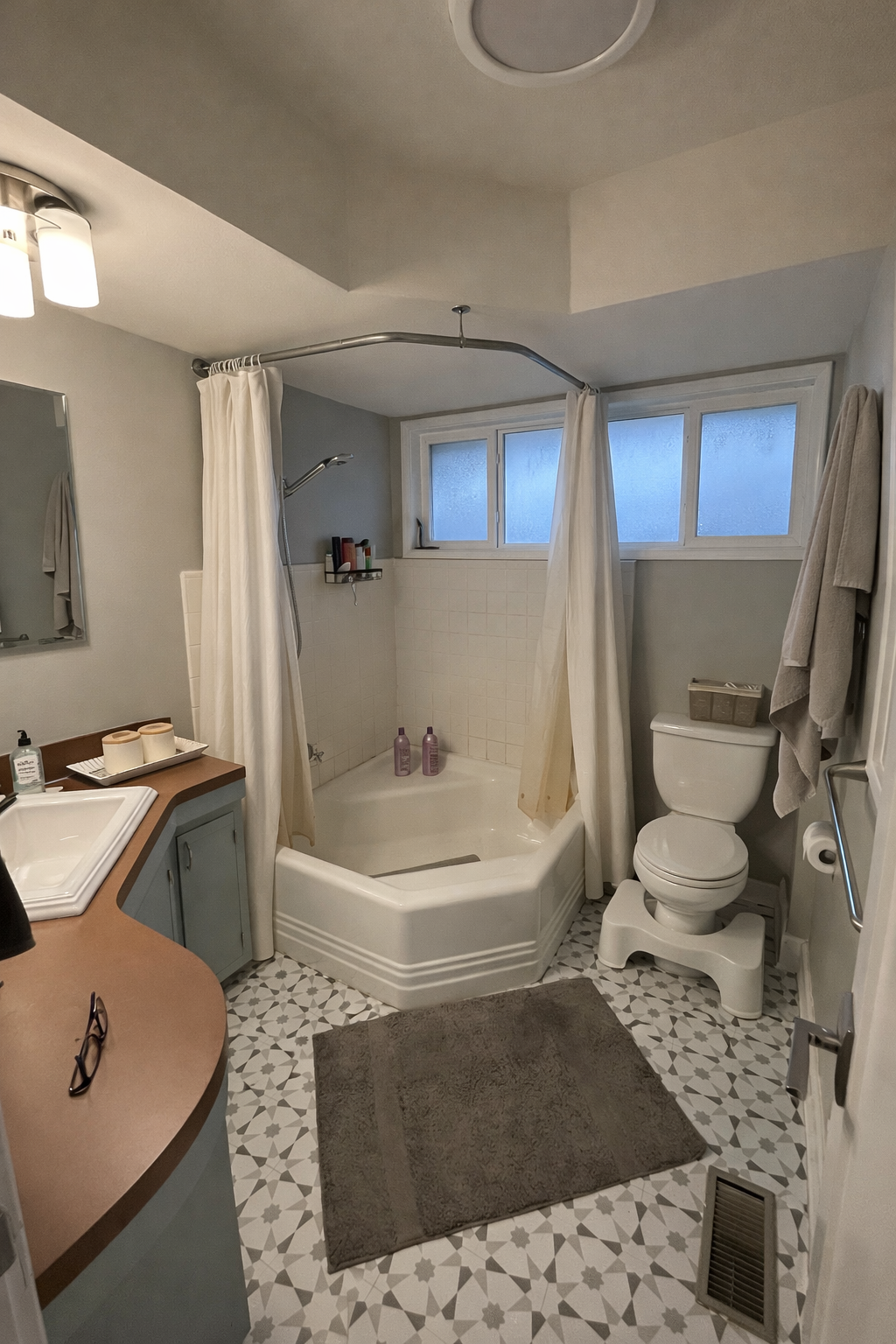A bathroom featuring a corner bathtub with white tiled walls, a window above, a toilet to the right with towels hanging on a rack, a gray bathroom mat on patterned floor tiles, and a vanity with sink and toiletries on the left.