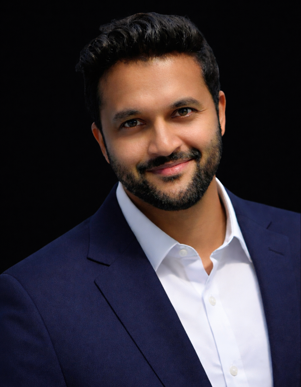 A professional headshot of a man with dark hair and beard, wearing a navy blazer and white shirt, smiling against a black background.
