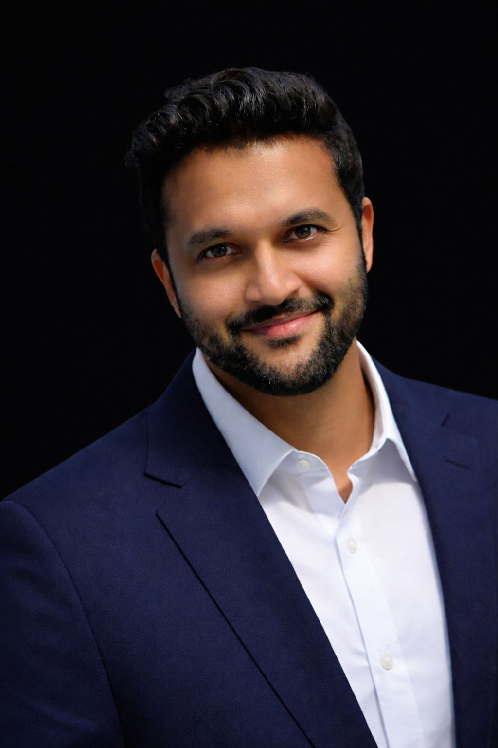 A man with dark hair and a beard, wearing a navy blazer and white shirt, smiling against a black background.