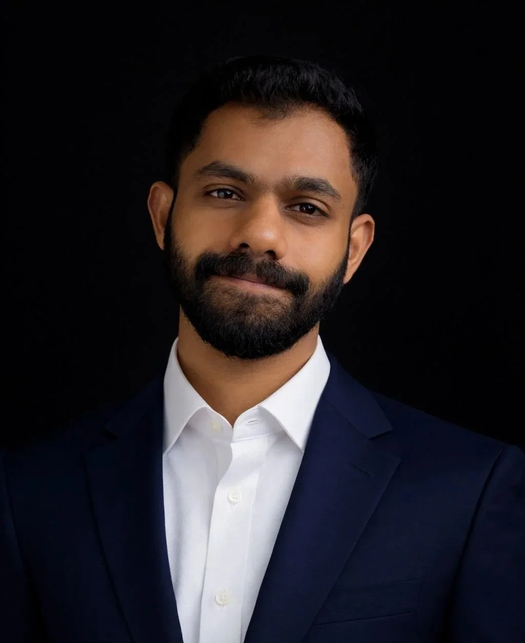 Headshot of a man with dark hair, a beard, dressed in a white shirt and dark blazer against a black background.