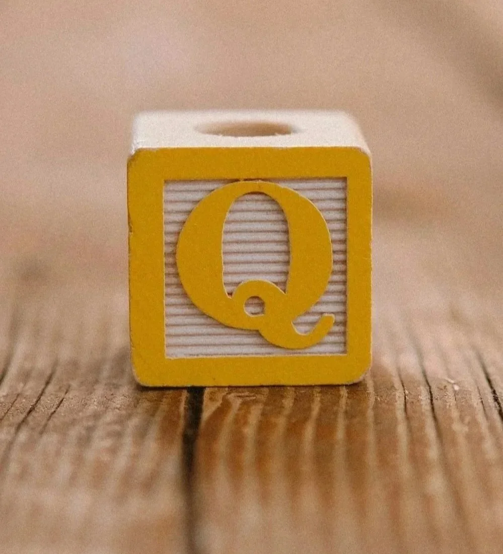 A small wooden block with a yellow border and white background featuring the letter 'Q' in yellow, placed on a wooden surface.