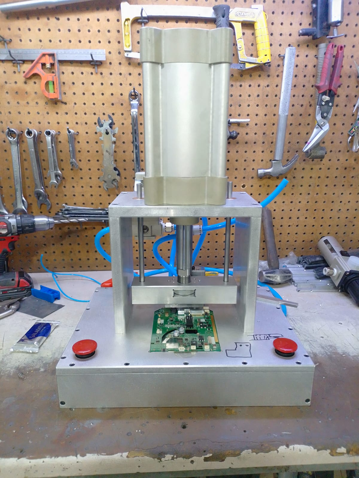 A workbench with a piece of electronic equipment, possibly a testing device, featuring a green circuit board and red buttons. The background has a pegboard with various hand tools like wrenches and saws hanging on it.