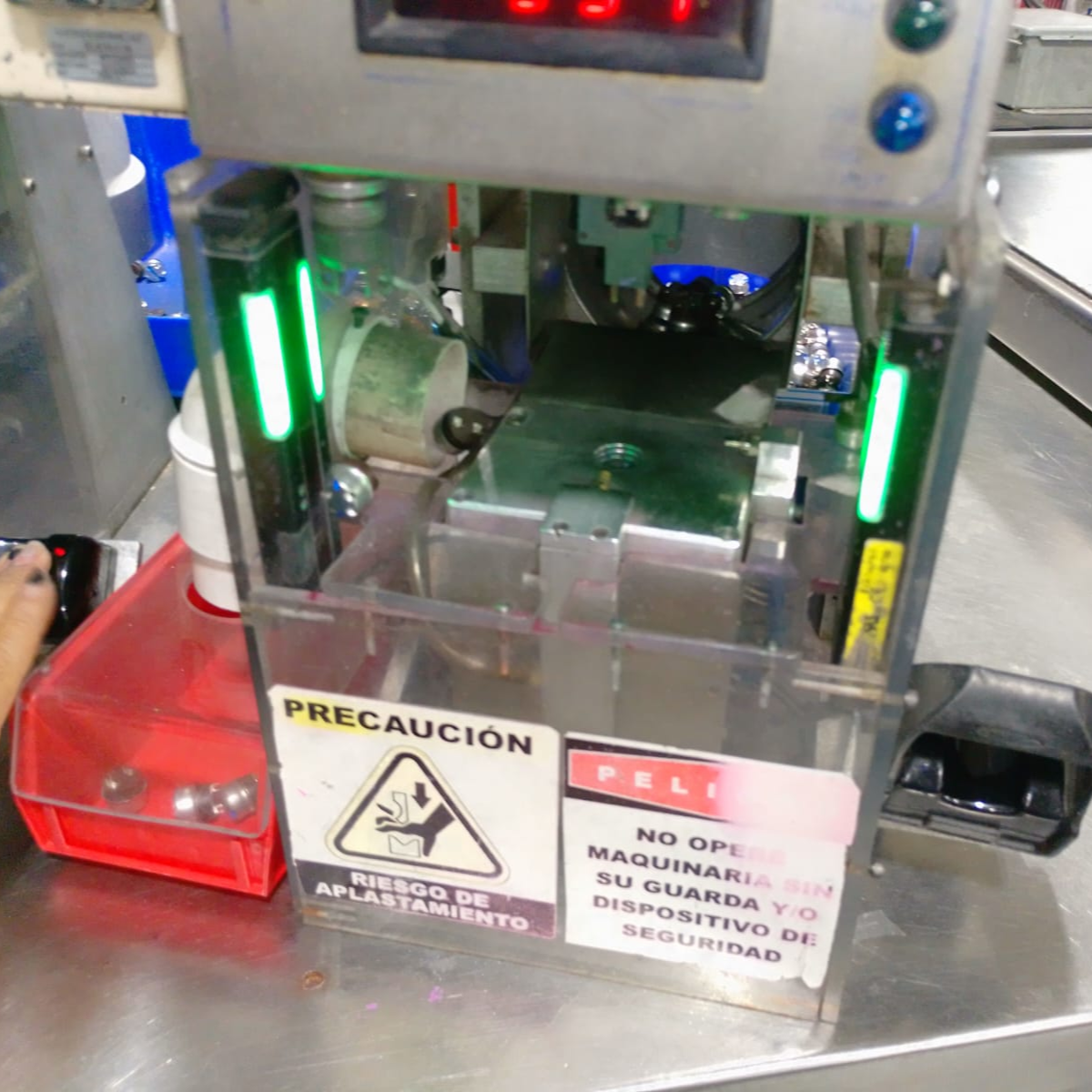Small industrial machine with warning labels, metal components, and green indicator lights, used for manufacturing or assembly processes.