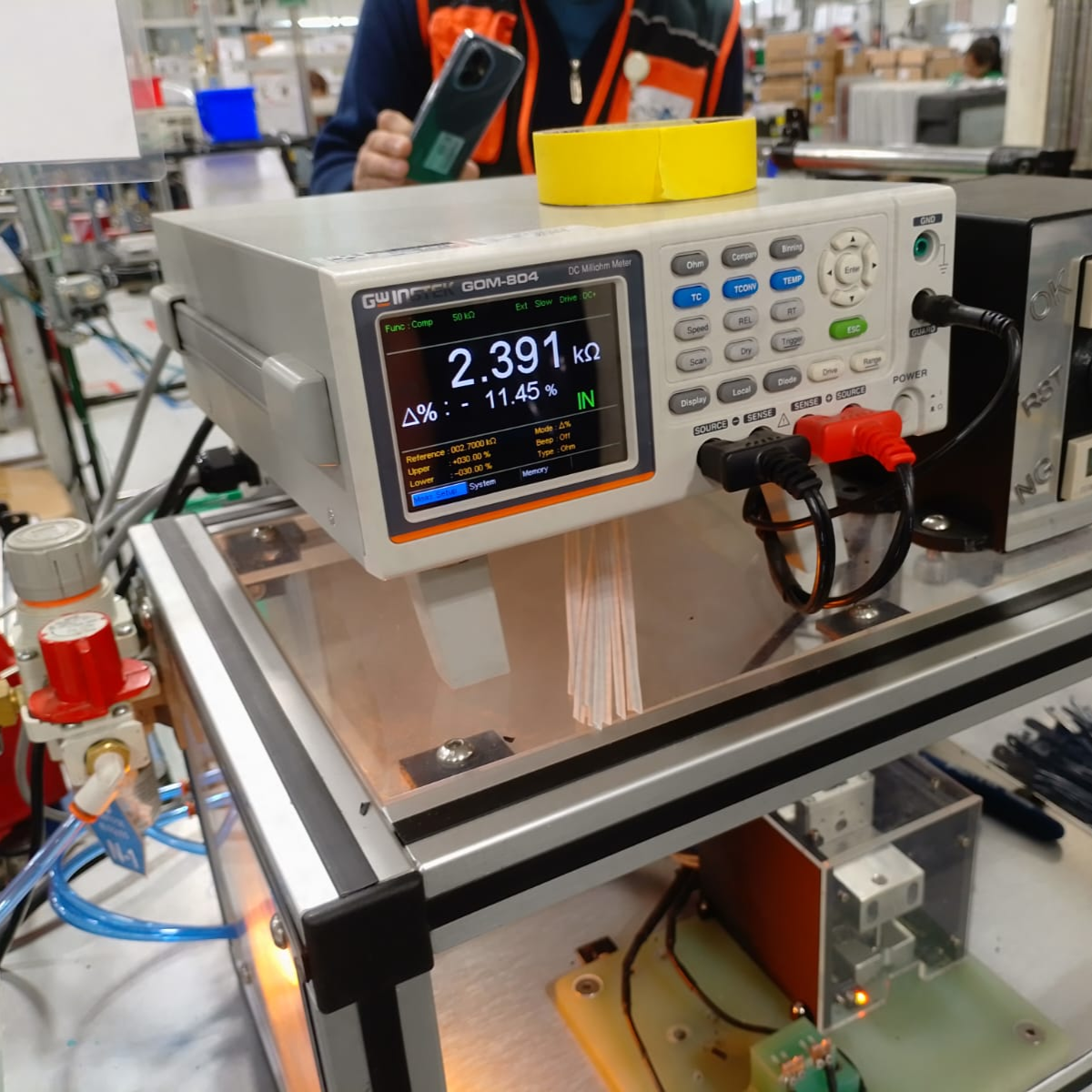A digital electronics testing device measuring resistance with a display showing 2.391 kilo-ohms and a person in the background holding a smartphone in a manufacturing or laboratory setting with electronic components and equipment visible.