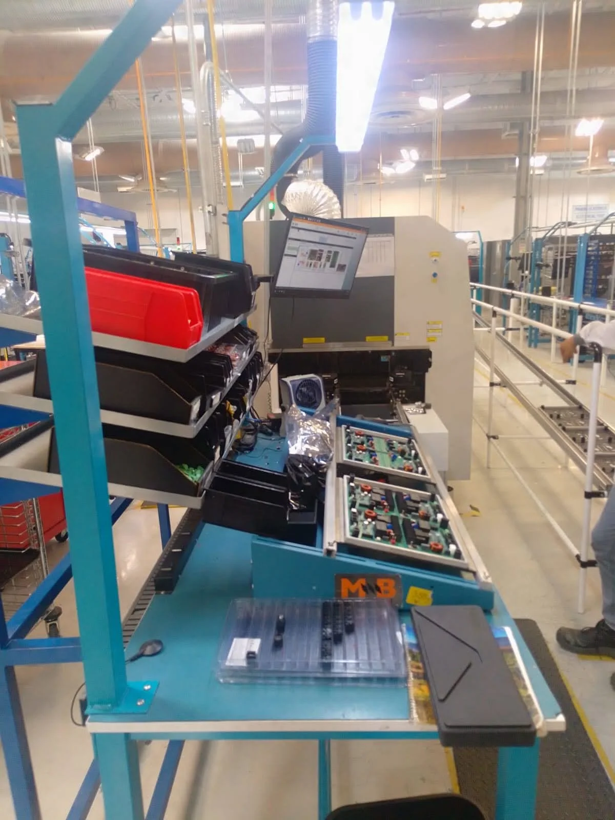 Factory workstation with electronic components and tools, a computer monitor, colorful plastic bins, and assembly equipment in an industrial setting.