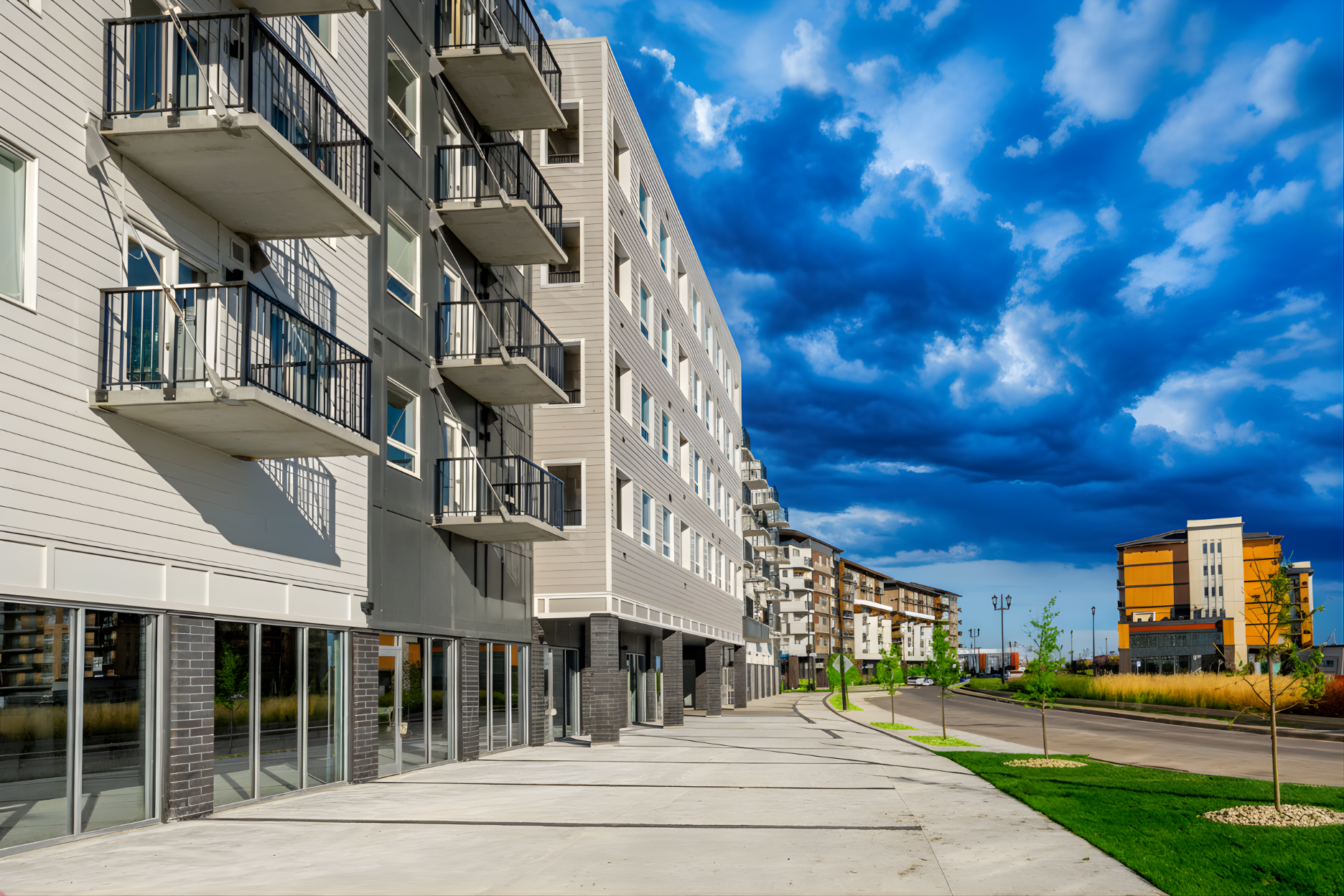 Modern multi-story apartment buildings with balconies, a spacious sidewalk, and green landscaping in an urban area under a blue sky with clouds.