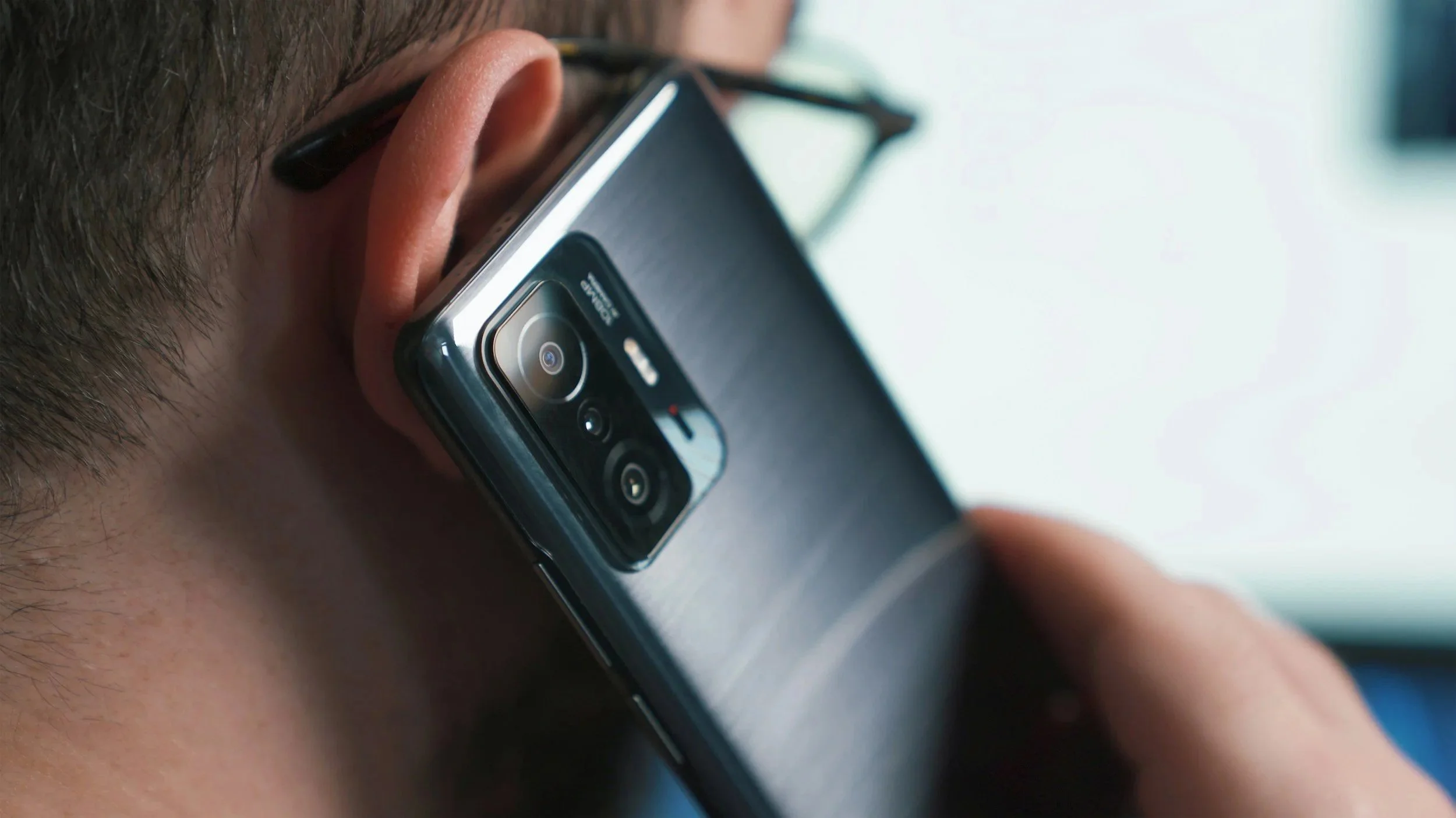 A person holding a smartphone at an angle, showing the back camera lenses and part of the person's face.