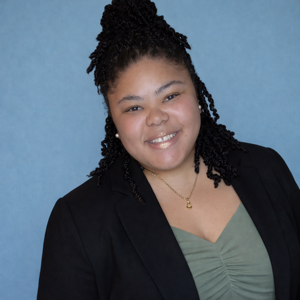 A woman with dark, curly hair styled in twists, wearing a black blazer and an olive green top, smiling against a blue background.