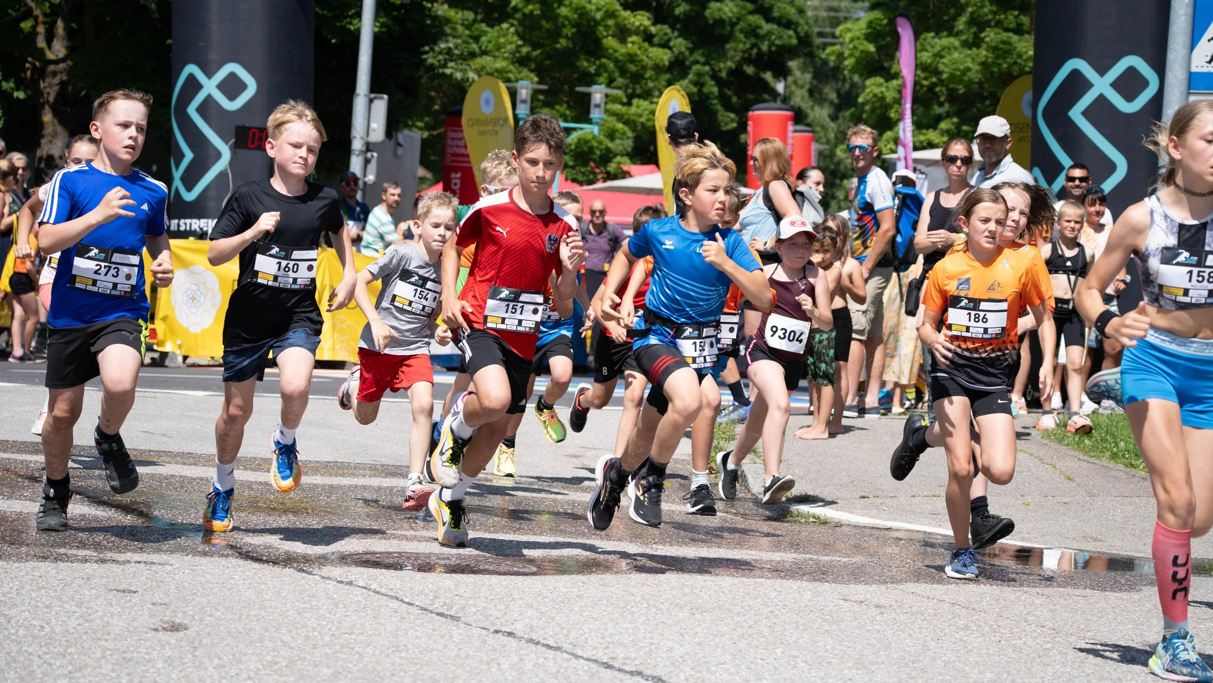 Kinder laufen in einem Wettrennen auf einer Straße, Hintergrund mit Zuschauern, bunten Bannern und grünen Bäumen.