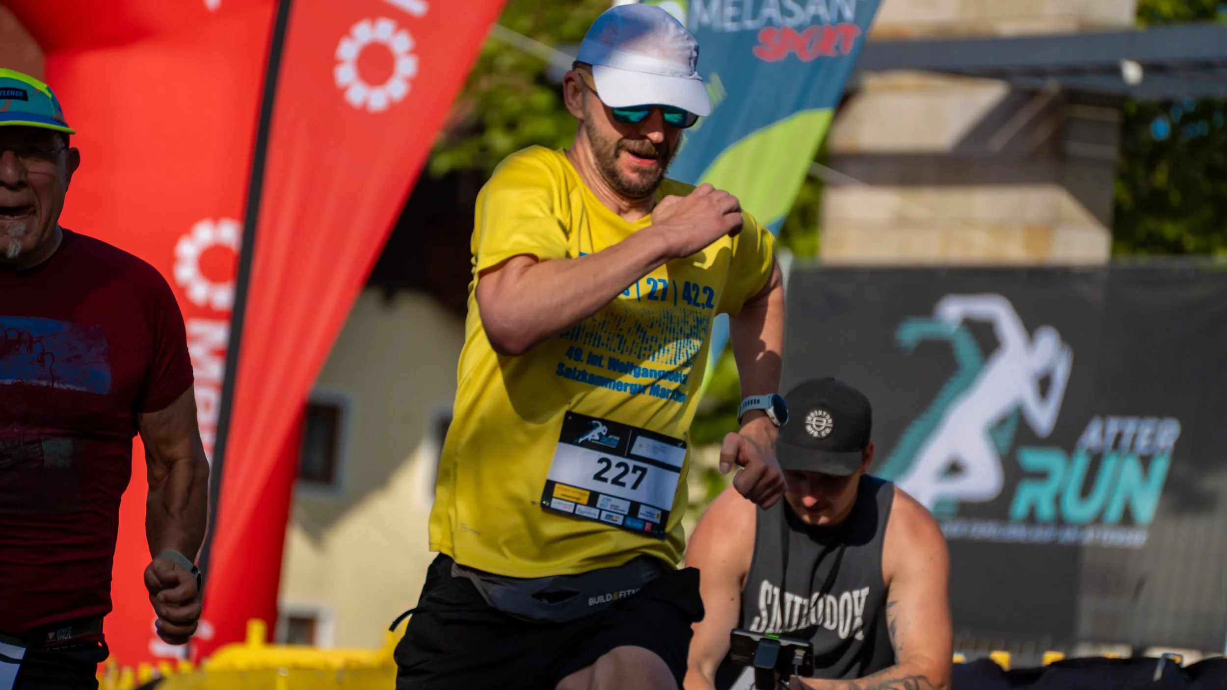 Mann im gelben Laufshirt läuft bei einem Wettkampf, im Hintergrund weitere Läufer und ein Banner mit der Aufschrift 'After Run'.