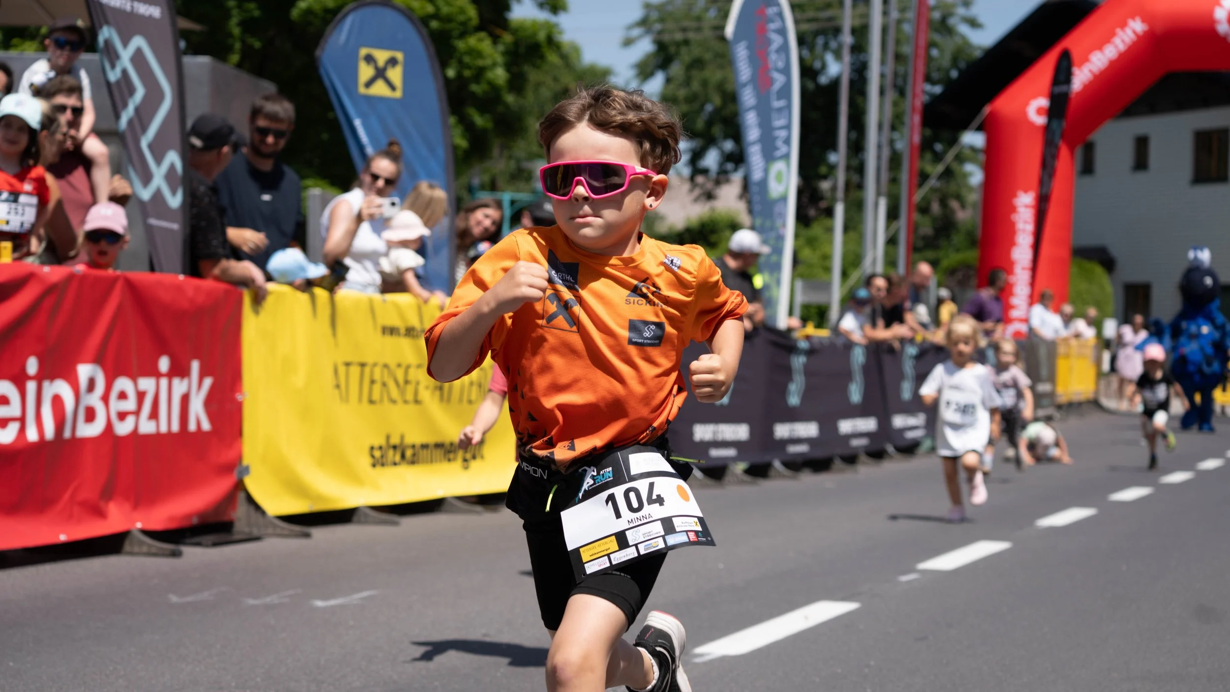 Junge beim Kinderlauf in orangeem Shirt und pinker Sonnenbrille läuft auf Straße, im Hintergrund stehen und sitzen Zuschauer hinter Absperrungen.