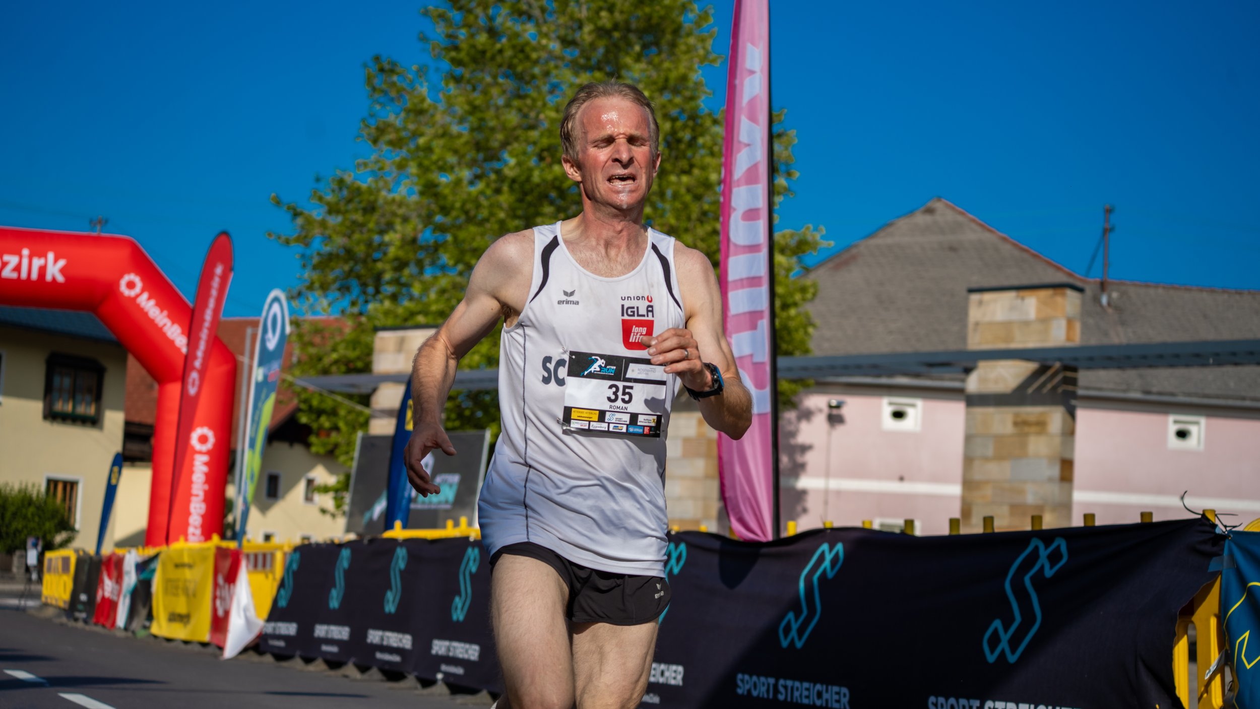 Mann beim Laufen an einem Straßenrennen, trägt einen weißen Laufshirt mit Nummer 35, im Hintergrund bunte Flaggen und Bauten bei sonnigem Himmel.