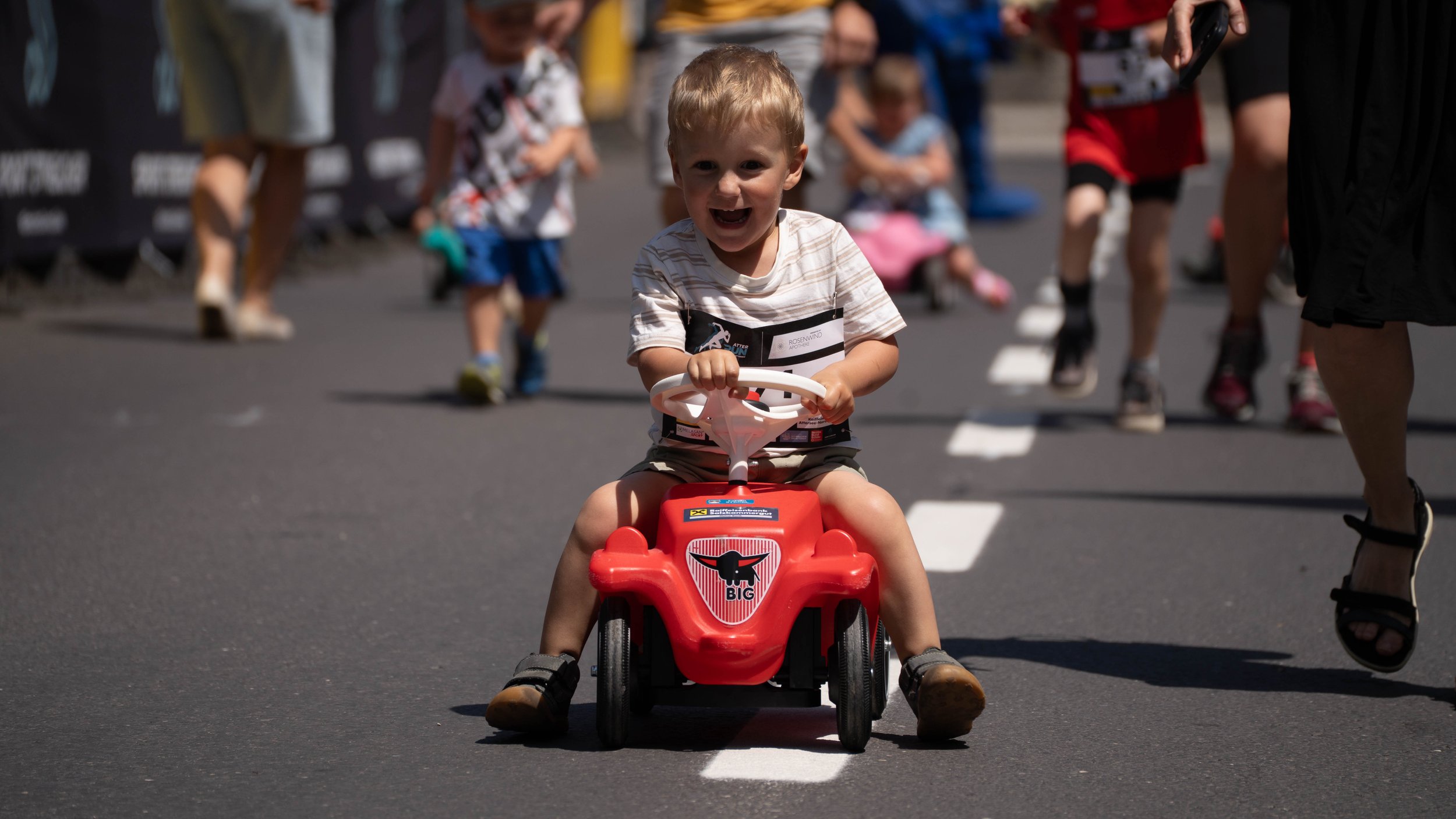 Ein lachender Junge auf einem roten Spielzeugauto fährt auf einer Straße mit weißen Linien, umgeben von mehreren Kindern und Erwachsenen.