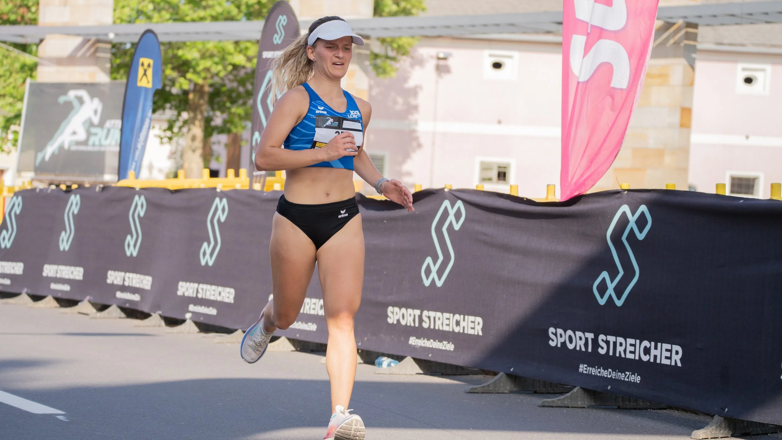Läuferin beim Laufrennen, trägt Sportkleidung und eine Visor, läuft auf der Straße neben einer Barriere mit Werbung.