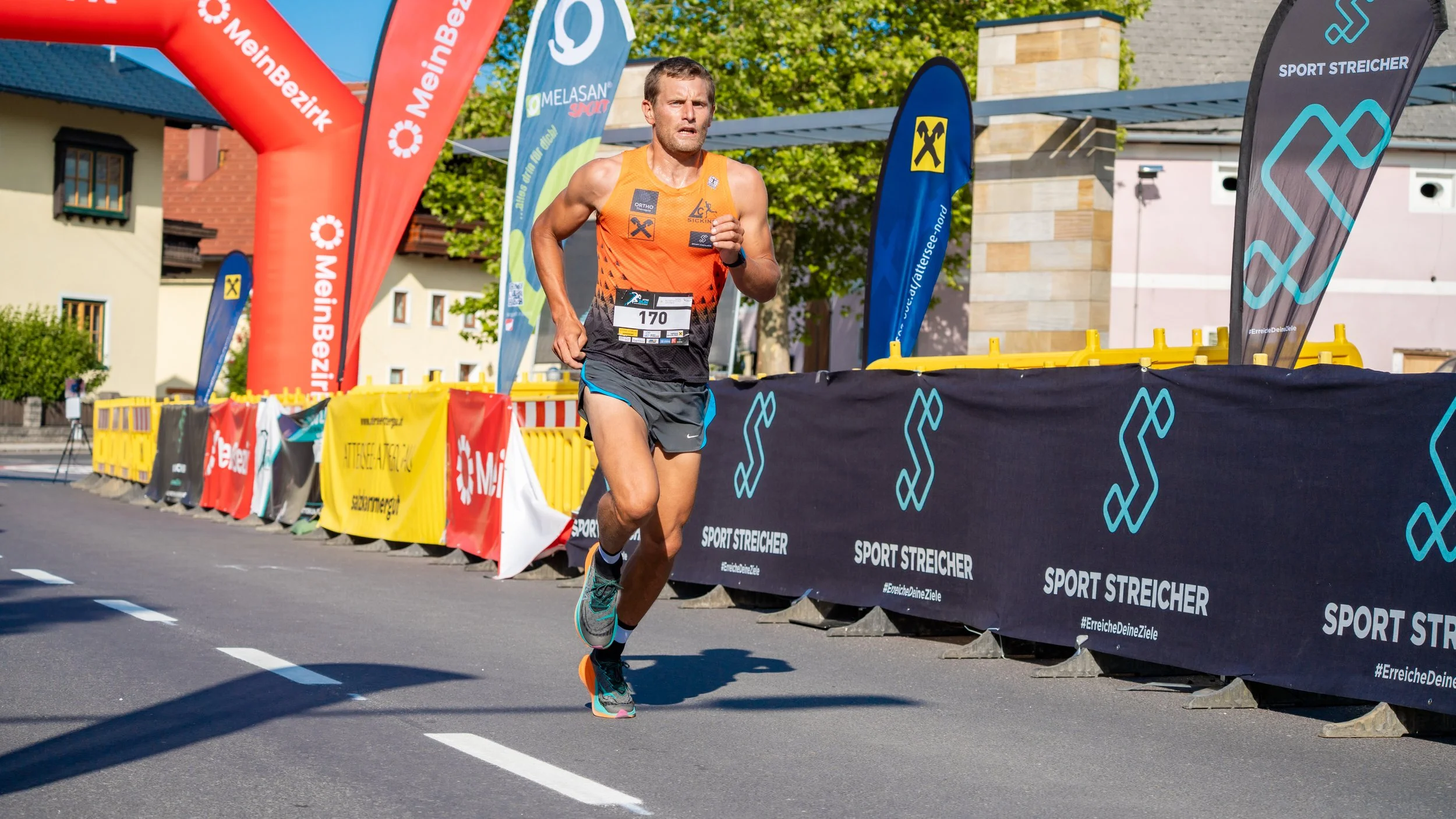Mann läuft bei einem Straßenlaufwettbewerb, trägt einen orangefarbenen Laufshirt und schwarze Shorts, neben Banden mit Sponsorenlogos, bei sonnigem Wetter mit blauen Himmel.