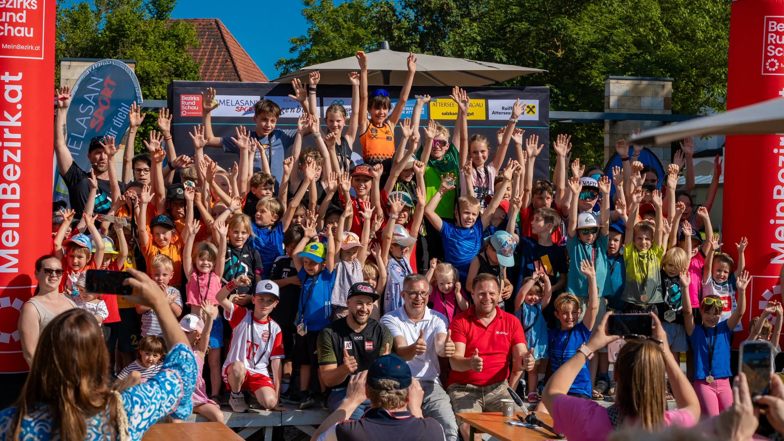 Gruppenfoto von Kindern und Erwachsenen bei einer Outdoor-Veranstaltung, alle heben die Hände in die Luft, im Hintergrund Bäume und blaue Himmel, auf bunten Bannern und Schildern ist österreichischer Text sichtbar.