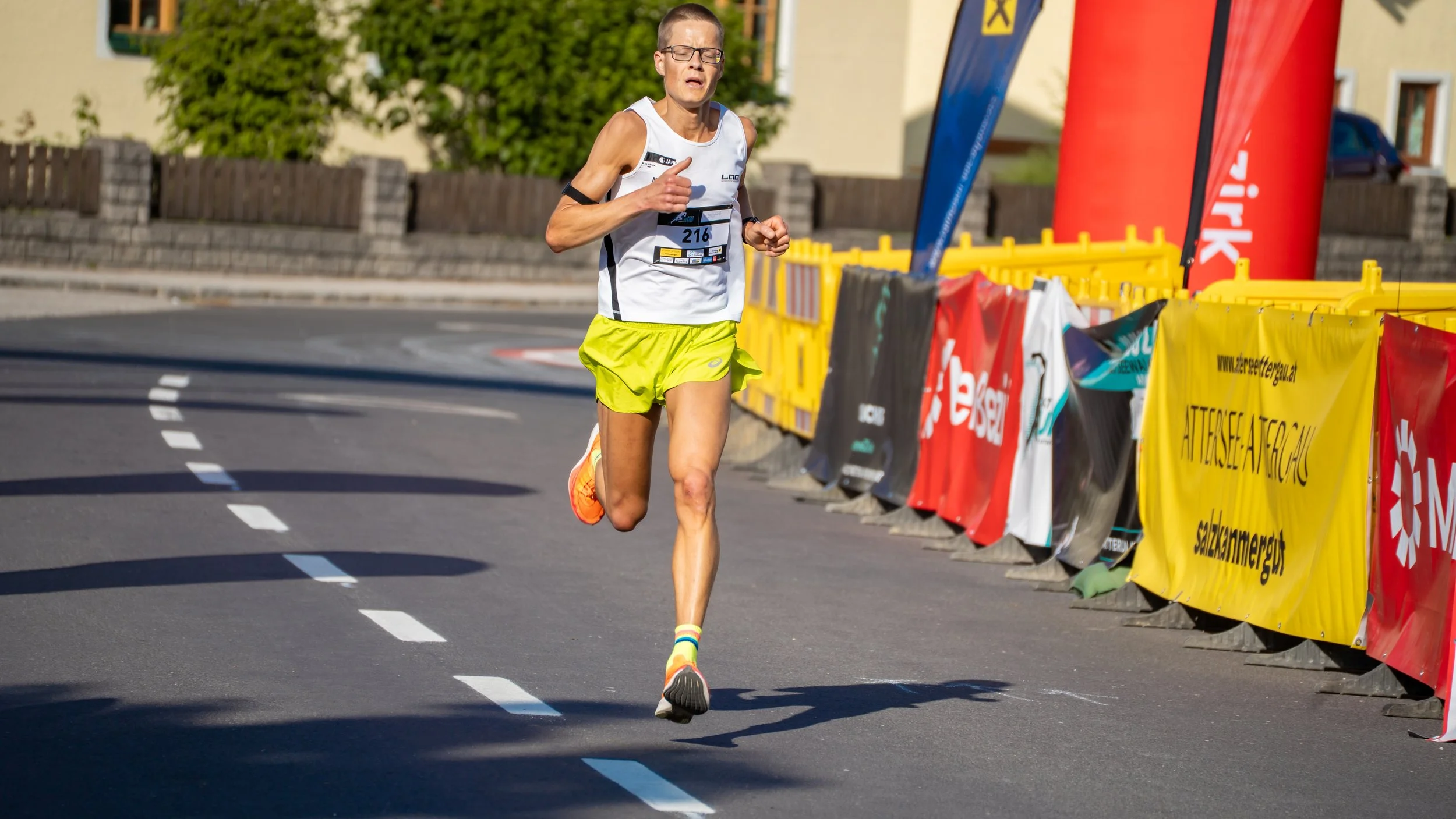 Ein männlicher Läufer beim Straßenmarathon, trägt weiße Tanktop und gelbe Shorts, läuft auf einer kurvigen Straße. Im Hintergrund sind Absperrungen mit bunten Bannern und Häuser zu sehen.