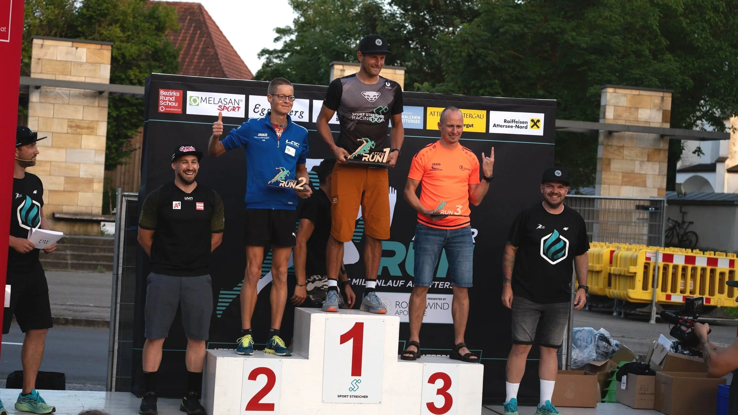 Siegerpodest bei einem Laufwettbewerb, mit drei Männern auf dem ersten, zweiten und dritten Platz, die Pokale in den Händen halten, umgeben von weiteren Personen. Im Hintergrund sind Bauten und Baken zu sehen.