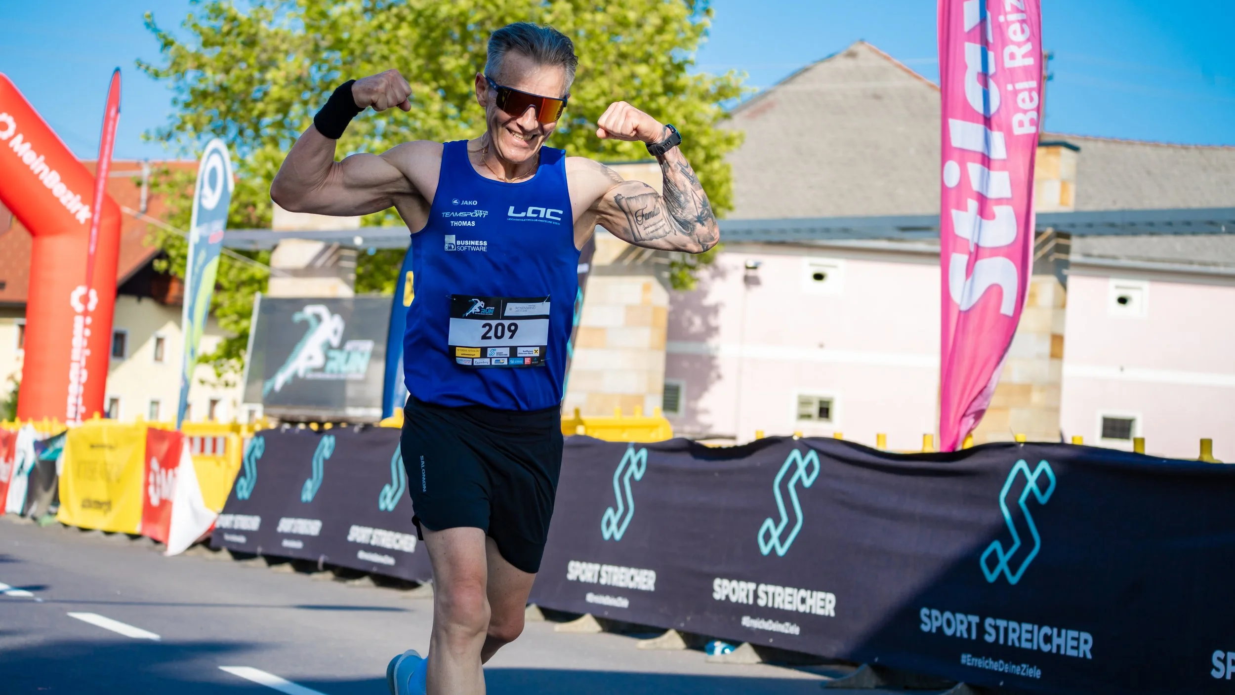 Ein männlicher Läufer zeigt seine Muskeln und lächelt, während er an einem Laufwettbewerb teilnimmt. Er trägt eine blaue Laufkleidung mit der Nummer 209, Sonnenbrille und schreibt auf seine Arme Tattoos.