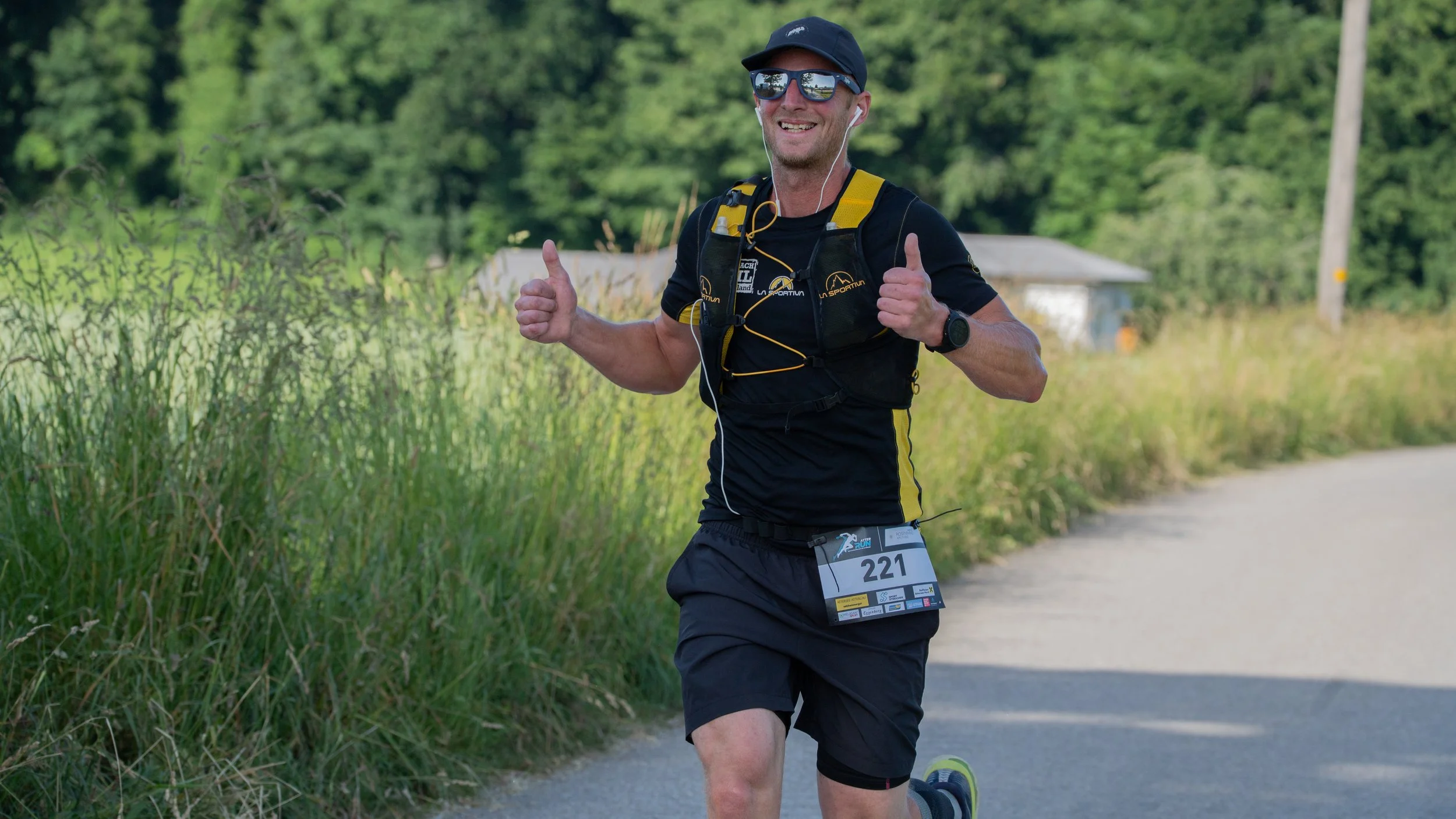 Ein Läufer bei einem Straßenlauf, trägt schwarze Kleidung, eine nummer 221, und lächelt mit Daumen hoch, während er auf einem Weg läuft, umgeben von Grün und Bäumen.
