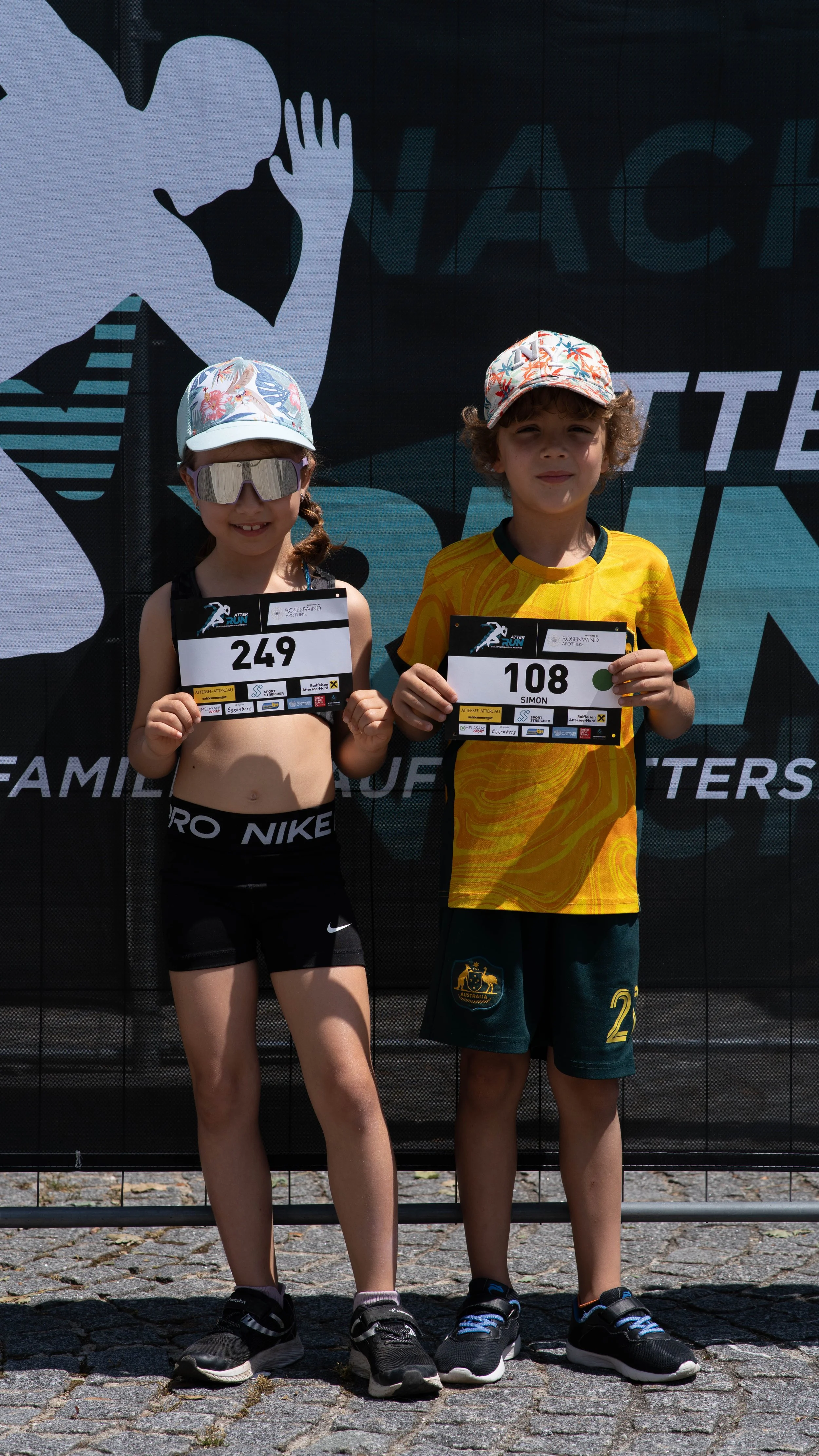 Zwei Kinder mit Startnummern bei einem Lauf, die vor einem Banner stehen.