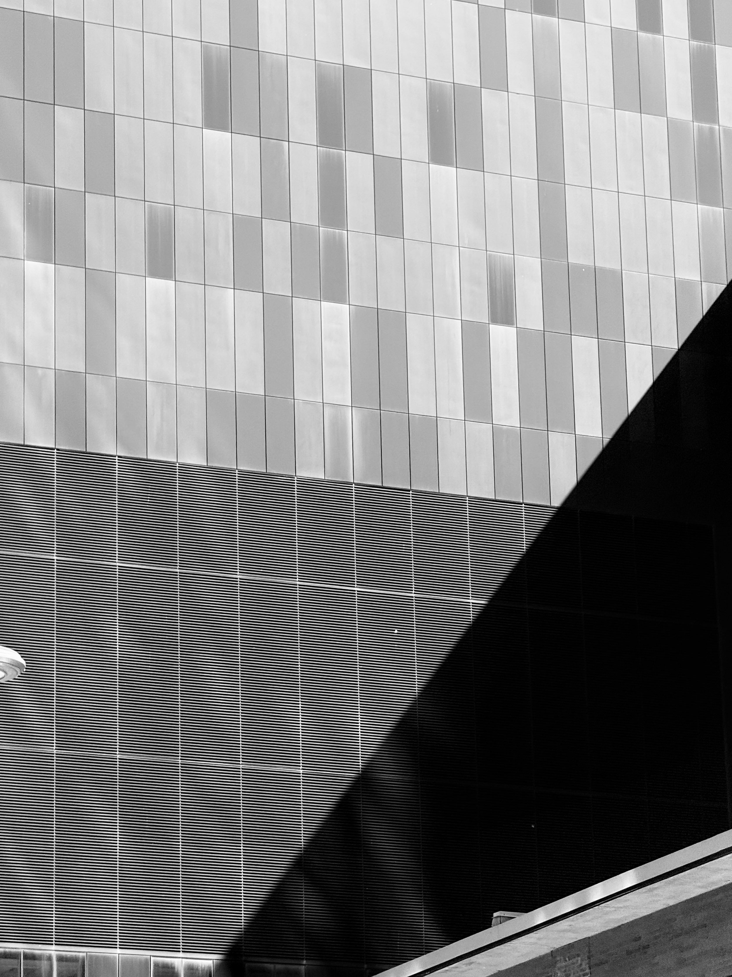 Black and white photo of a modern building with a curved, cladded facade and horizontal striped pattern. A portion of the building has small, square tiles, and there are architectural details such as a cylindrical object on the left and a building ledge at the bottom right.