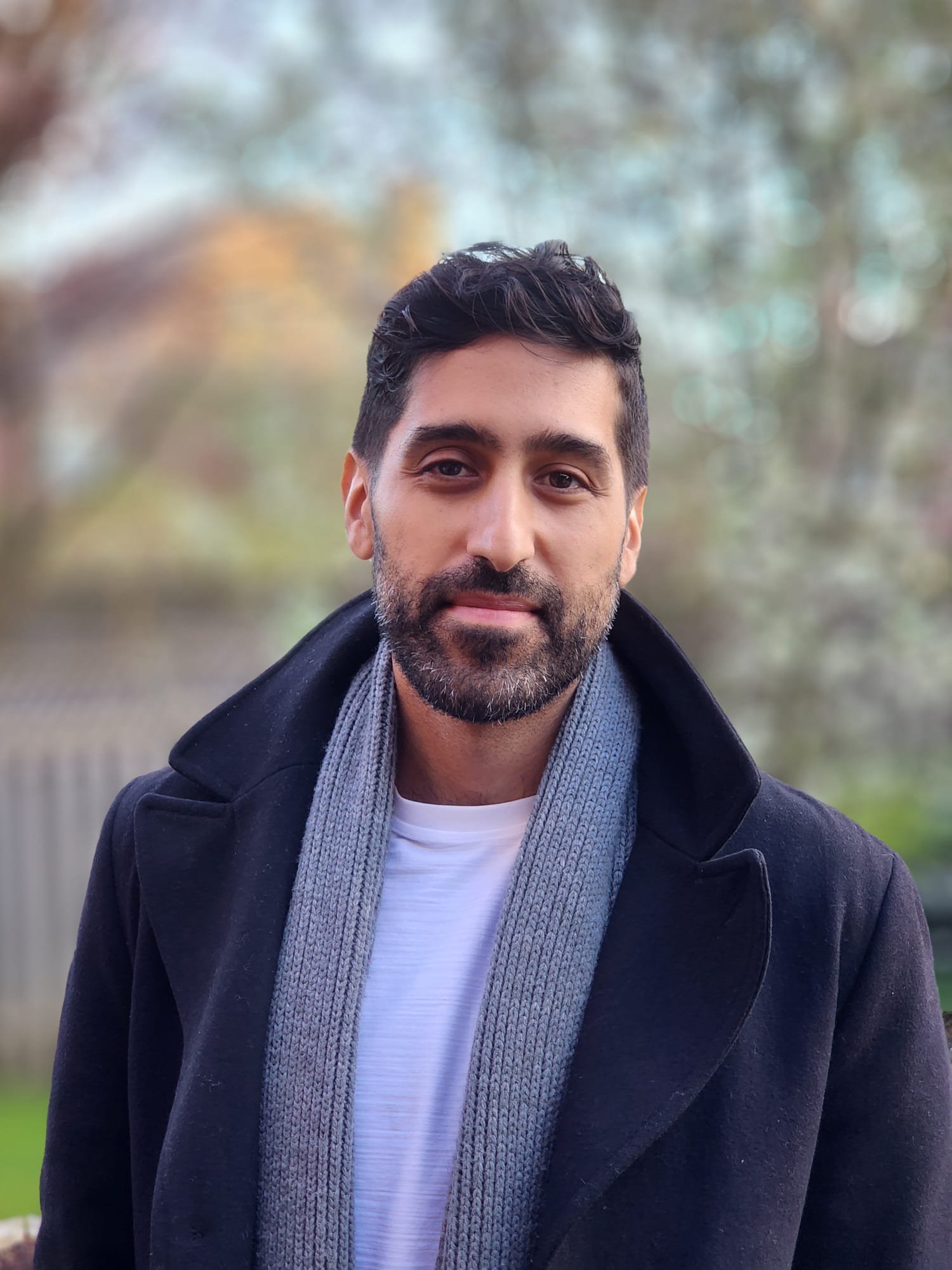 Close-up of a man with dark hair and a beard, wearing a black coat, gray scarf, and white t-shirt, outdoors during daytime, with a blurred background of trees and sky.
