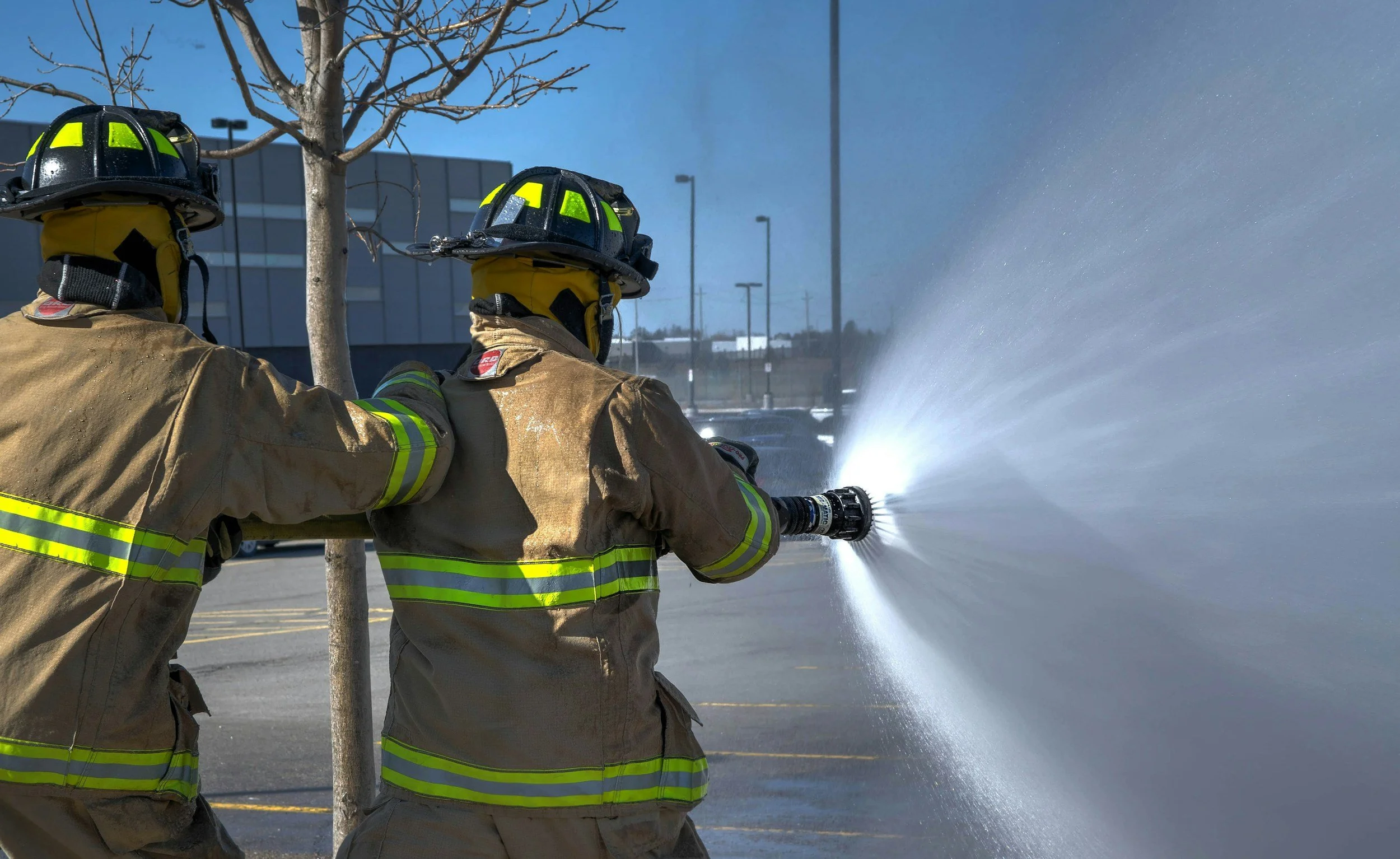 Two firefighters in full gear aiming a high-pressure water hose at a fire, with a building and parking lot in the background.