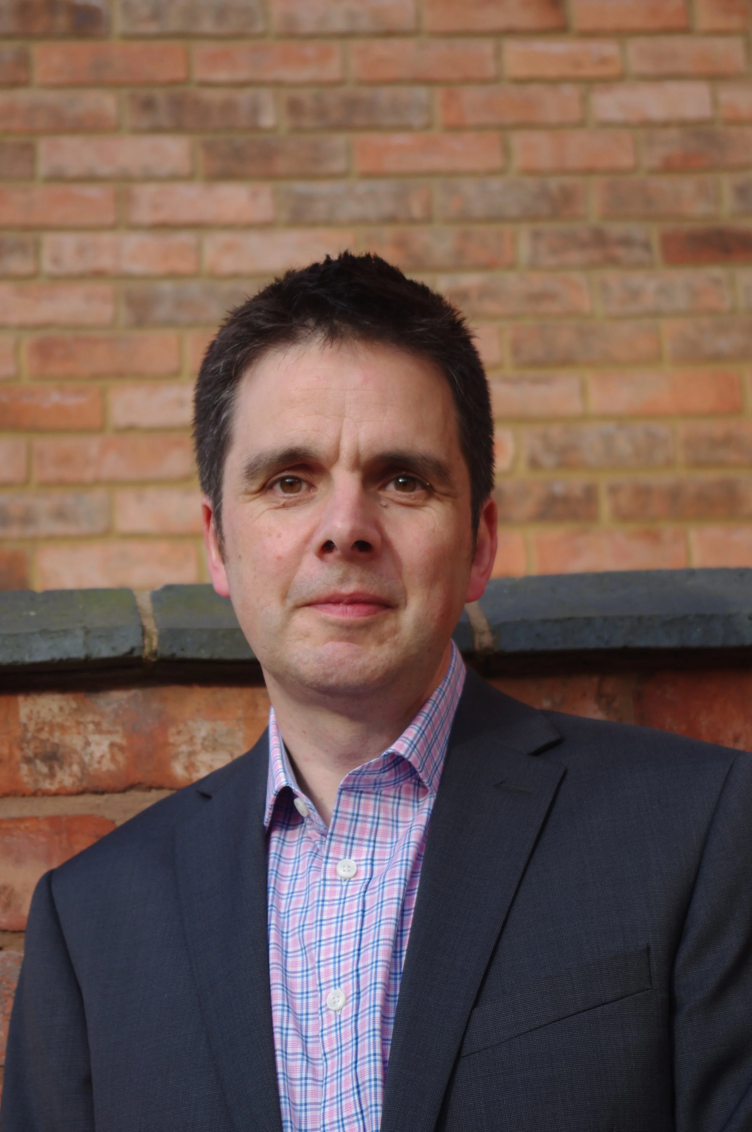 A man in a dark blazer and a pink checkered shirt stands in front of a brick wall.