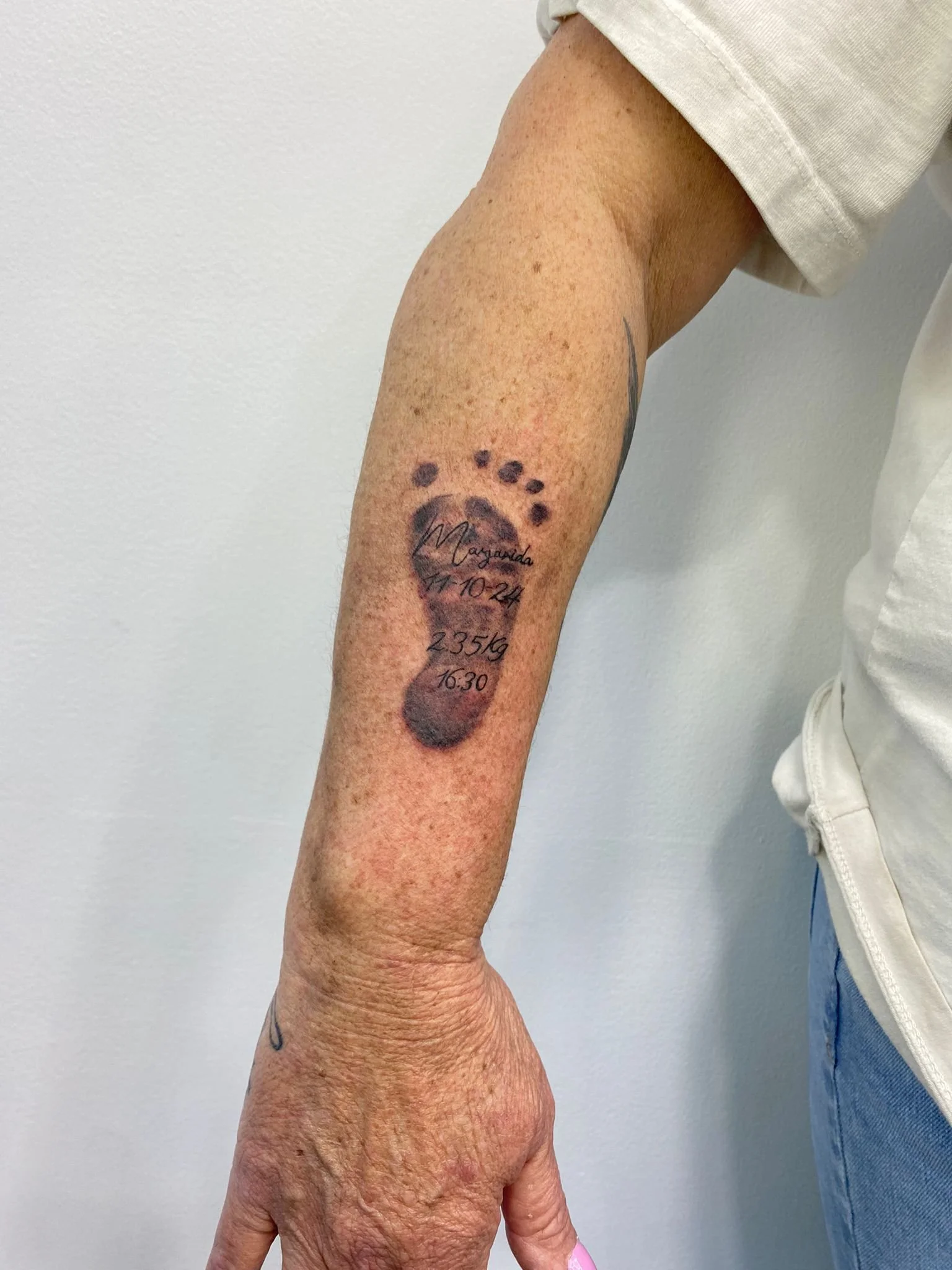 Tattoo de uma pegada com detalhes de datas, peso, nome e horário na parte interna do braço de uma pessoa.