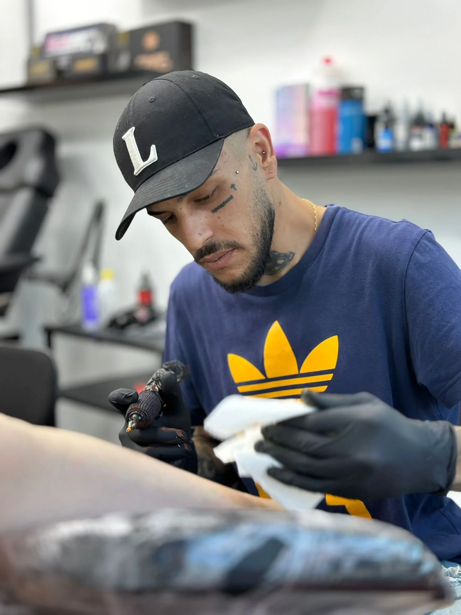João Câmara tatuando no estúdio de, usando uma camisa azul com o logo da Adidas, um boné preto com a letra L e usando luvas pretas.