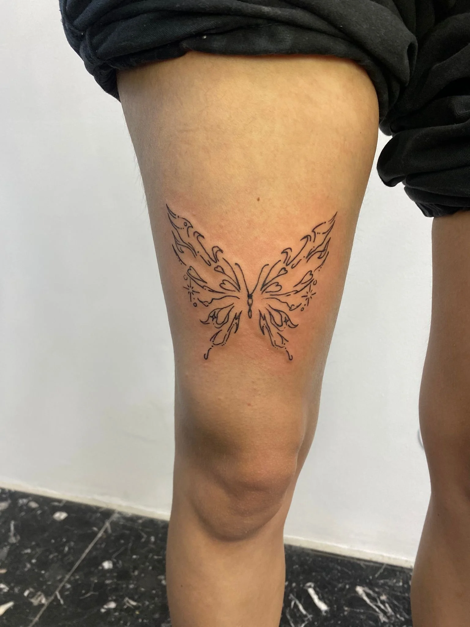 Tatuagem de uma borboleta com linhas detalhadas na parte frontal da coxa de uma pessoa, que está usando calças pretas.