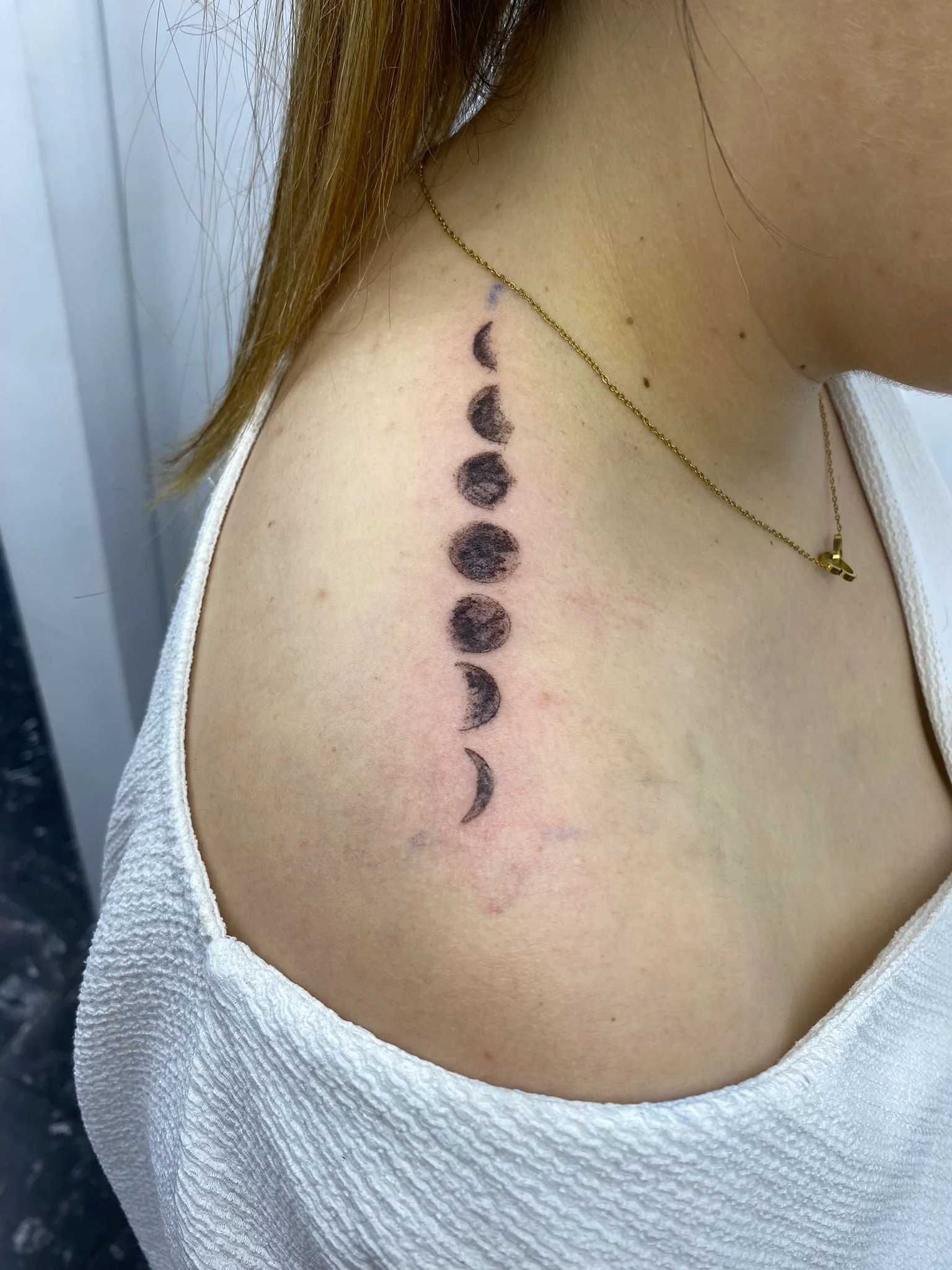 Tattoo de fases da lua na parte superior do braço de uma pessoa, com camiseta branca e usando colar dourado.