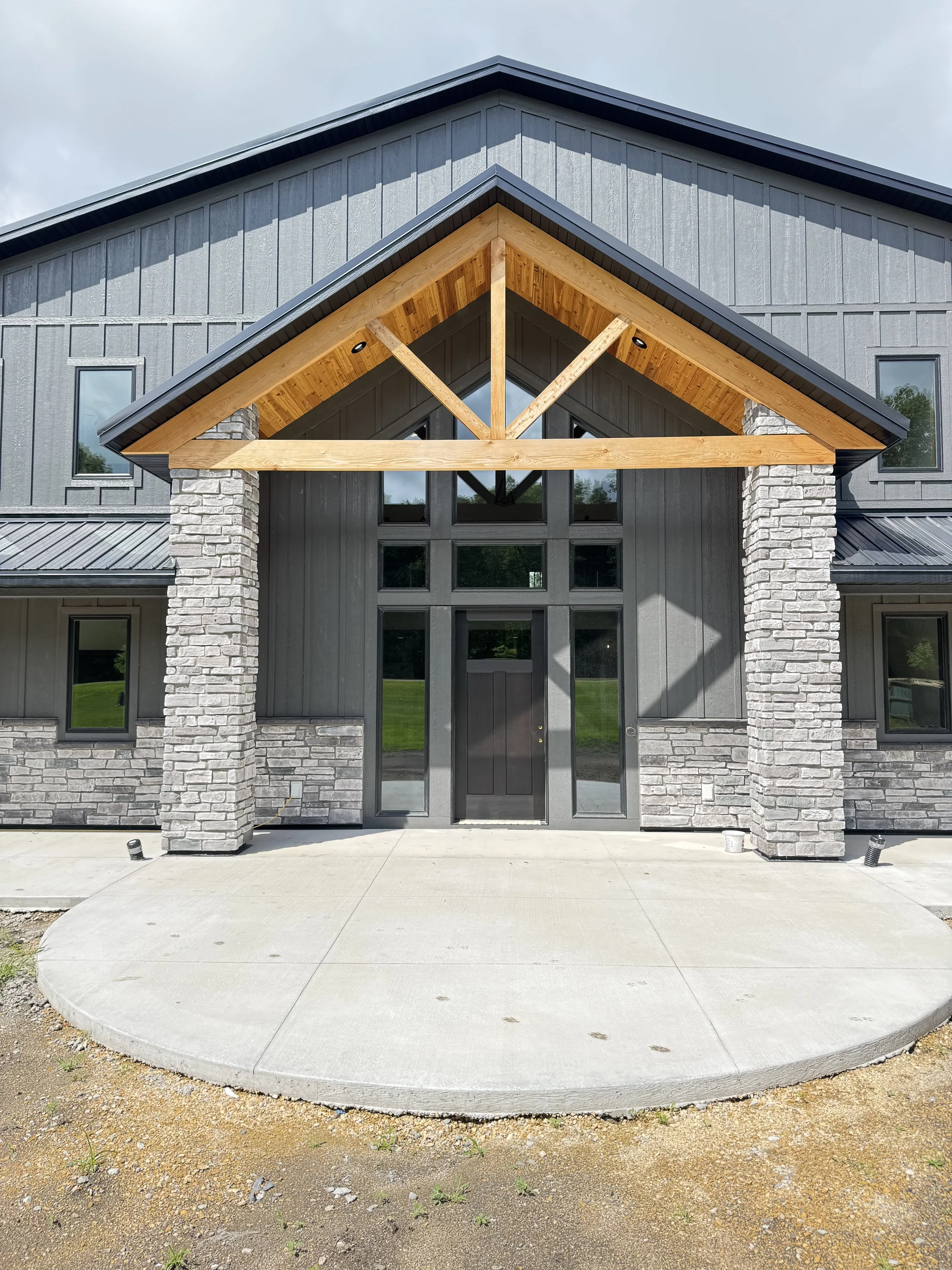 Front entrance of a modern house with a gabled roof, gray siding, brick columns, and large windows, with a concrete sidewalk and a small patch of dirt and grass in the foreground.