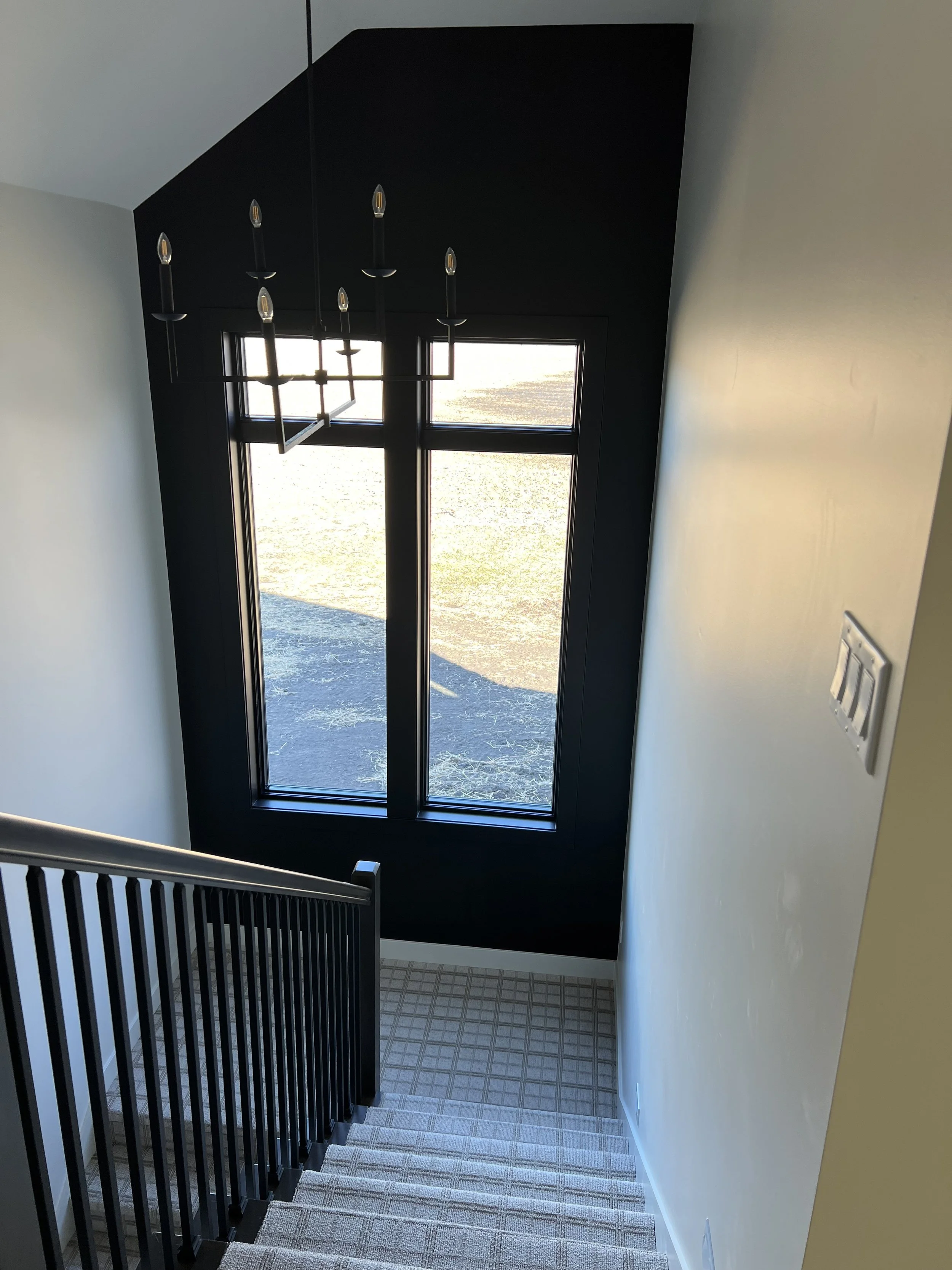 Modern staircase by Sprengeler Construction featuring black railing, large vertical windows, statement chandelier, and natural light-filled entryway