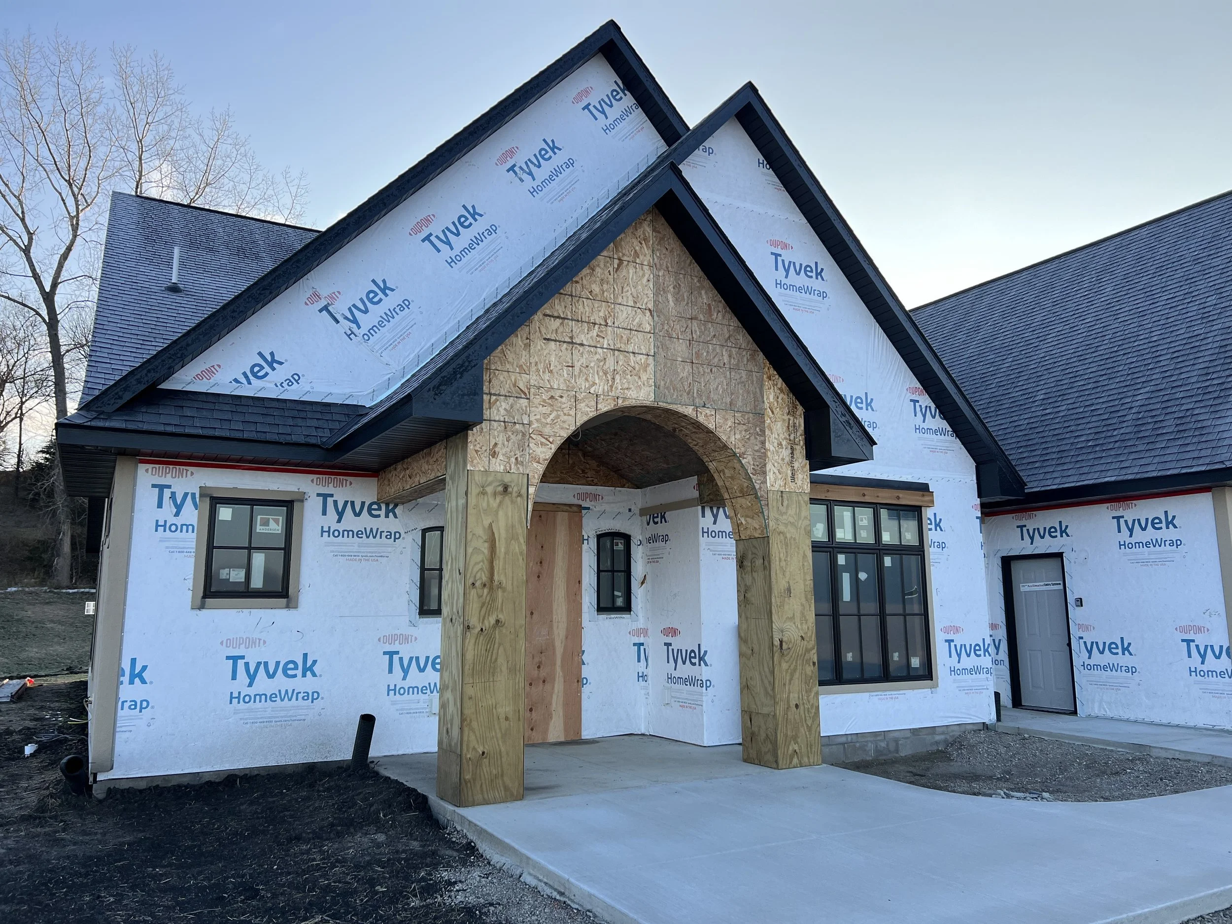 The image shows a house under construction with new framing and roofing. The exterior walls are covered with Tyvek HomeWrap, and the front entrance has a concrete porch with wooden posts but no door installed yet. The sky is clear and the surrounding area appears to be a suburban neighborhood.