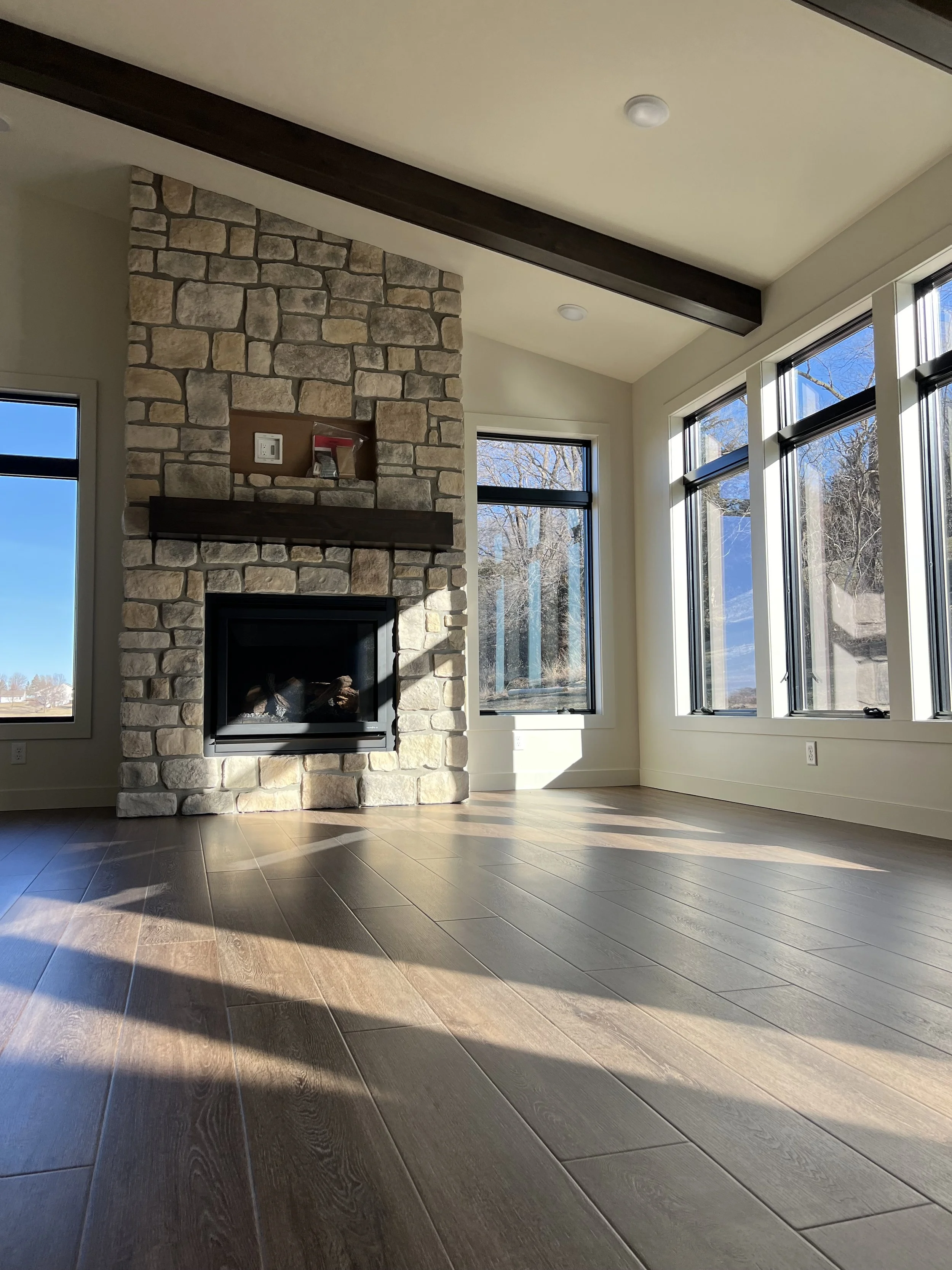 Living room by Sprengeler Construction featuring stone fireplace, exposed wood beam, hardwood floors, and large windows with natural light