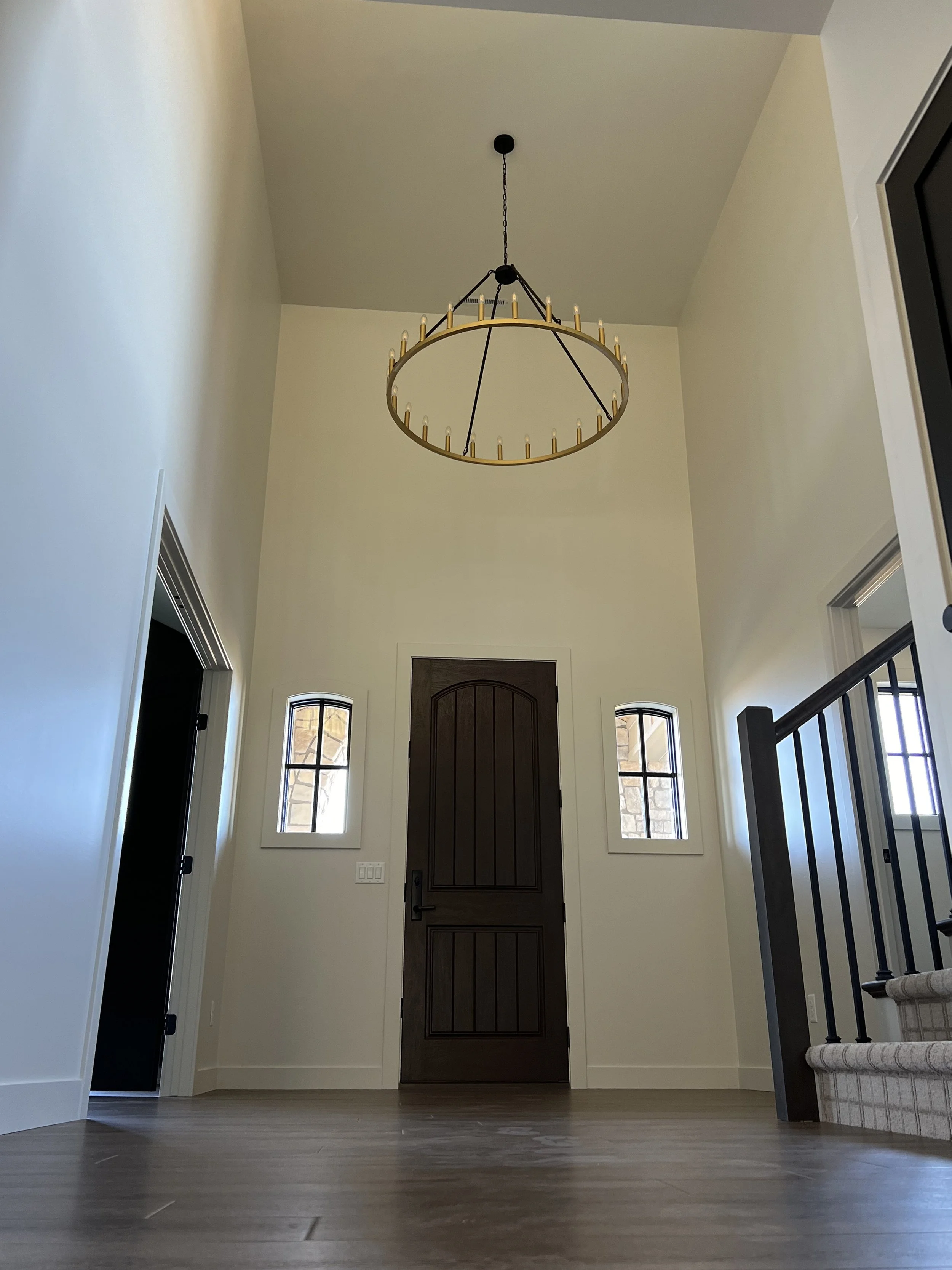 View of a house entryway with a dark wooden front door, two narrow windows flanking it, a staircase with a carpeted runner, and a large circular chandelier hanging from the ceiling.