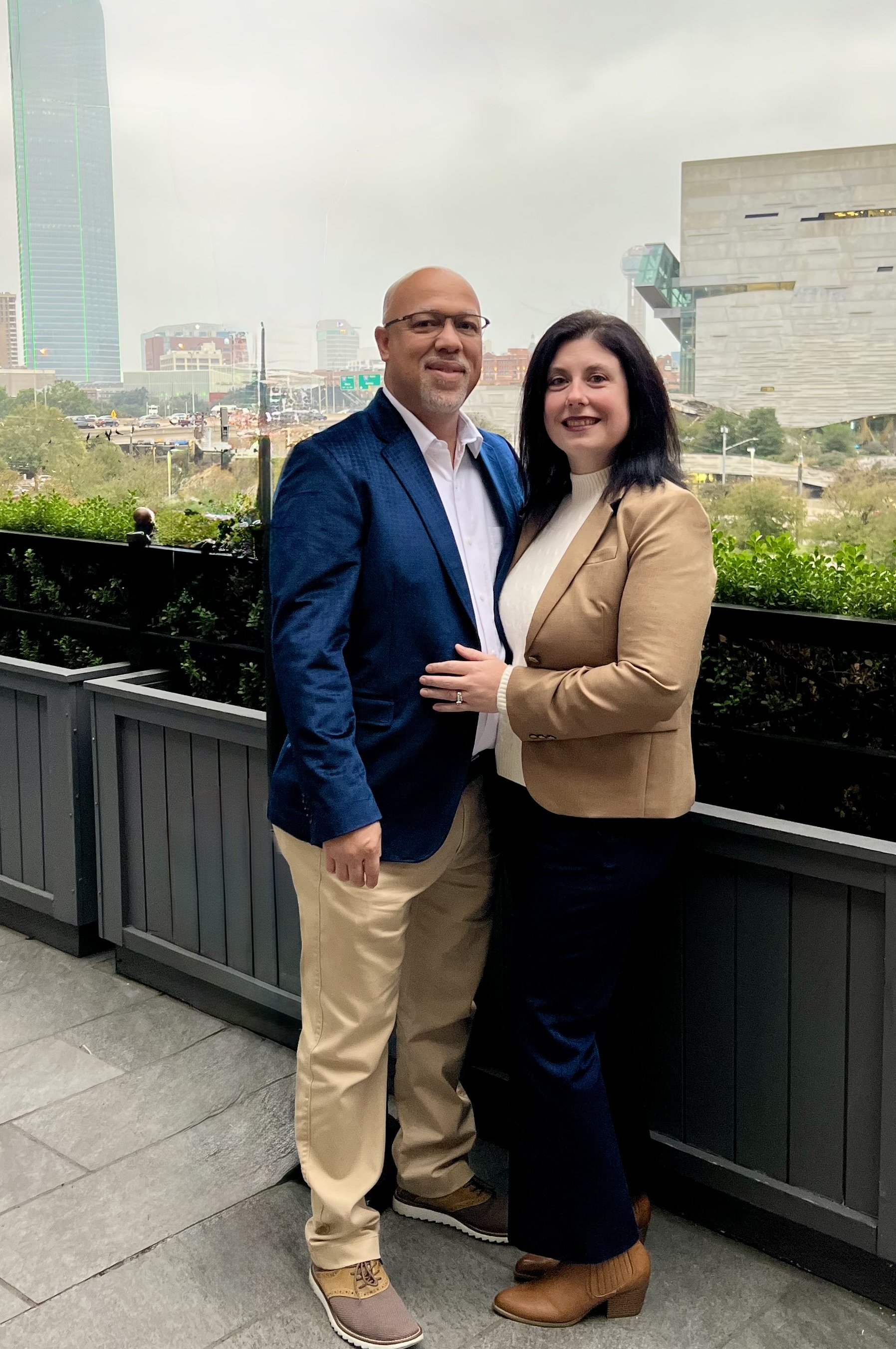 A man and woman standing together outdoors in front of a cityscape, smiling, with tall buildings and a cloudy sky in the background.