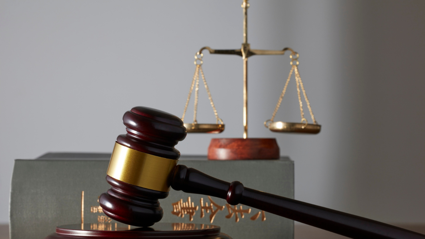 A wooden gavel resting on a sound block in front of a legal book, with a brass balance scale in the background.